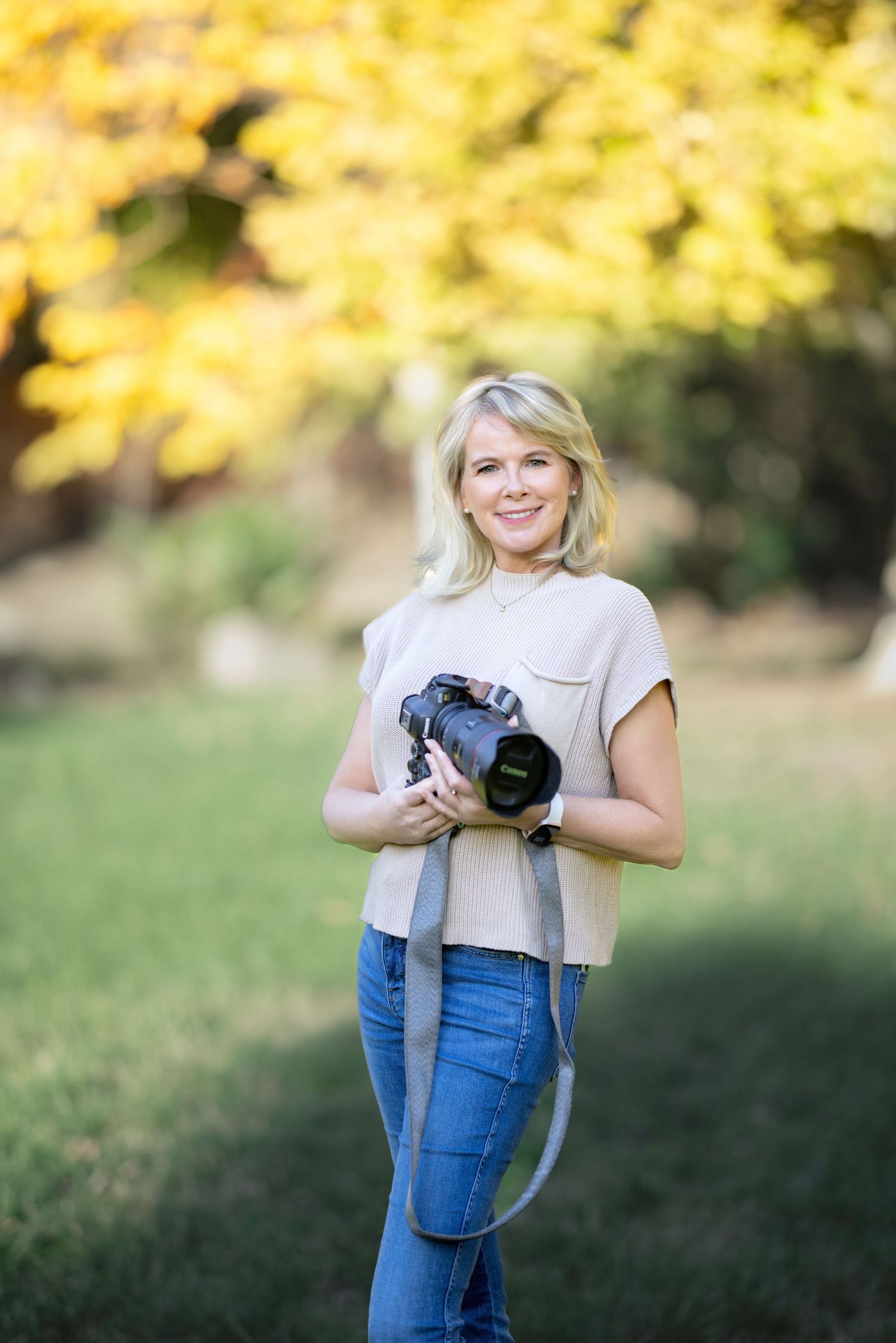 Professional photographer in casual attire holding a DSLR camera outdoors with autumn foliage in the background.