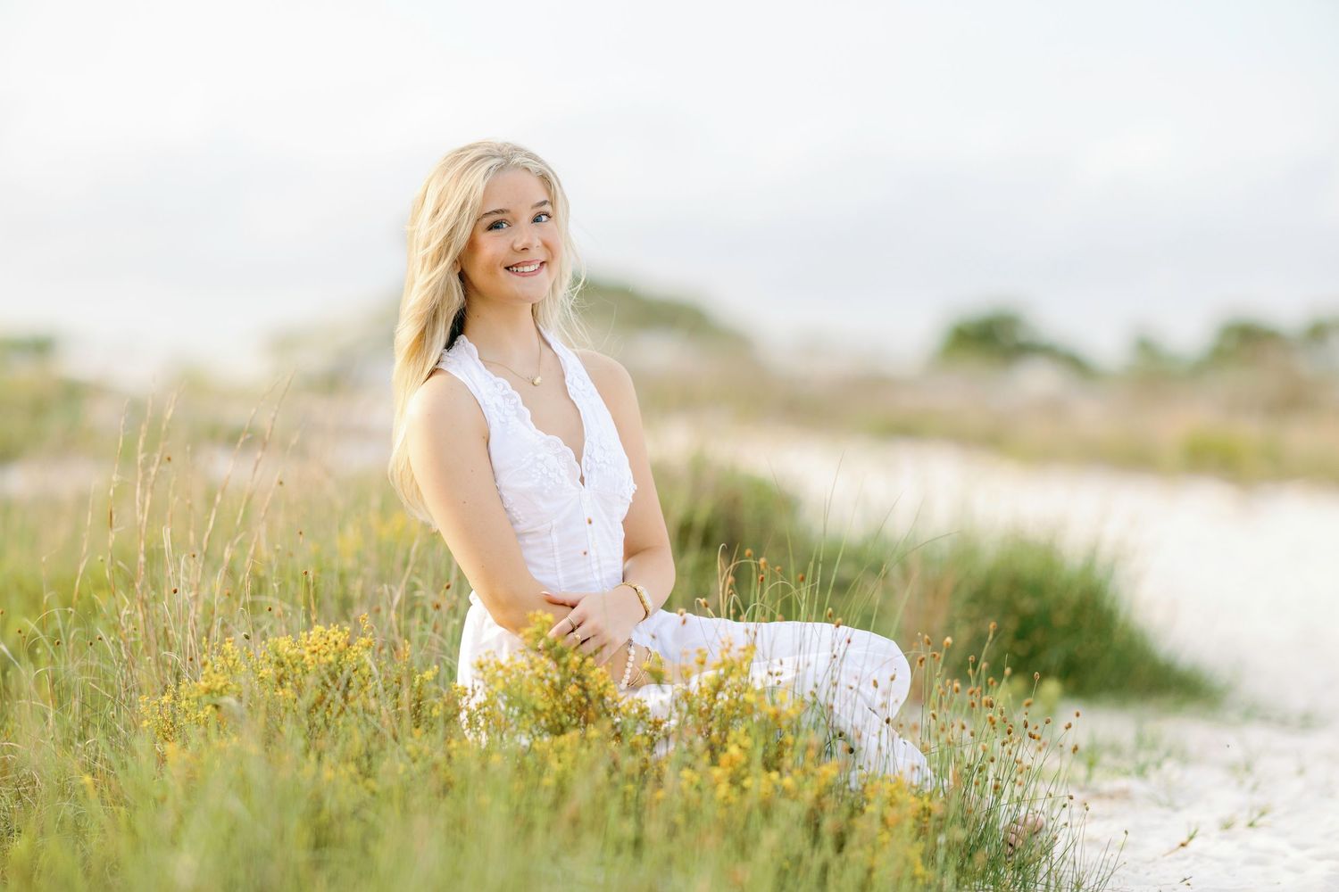 Woman in white dress sitting in field of yellow wildflowers on sunny day at beach.