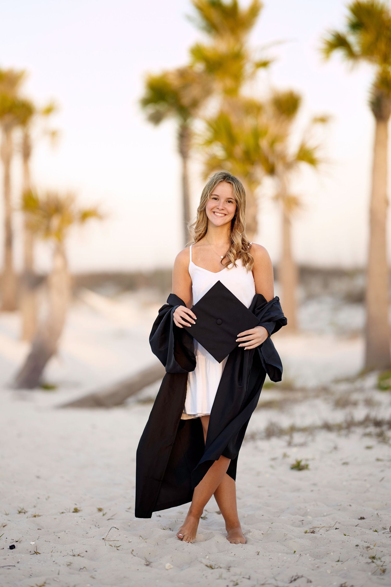 A woman in a black cardigan poses on a sandy beach with palm trees during sunset.
