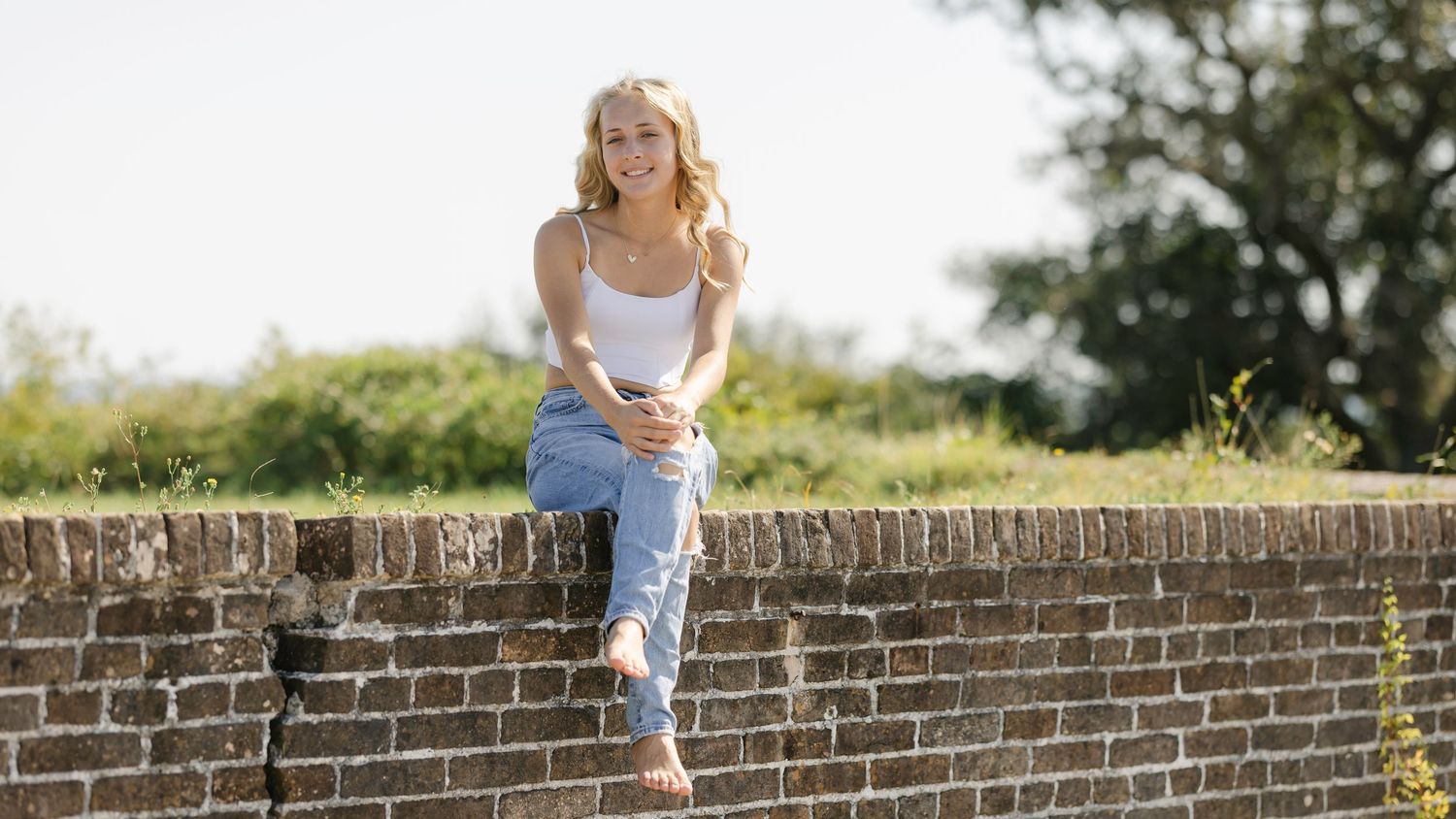 Young person in ripped jeans walking along a brick wall pathway in a summer garden setting.