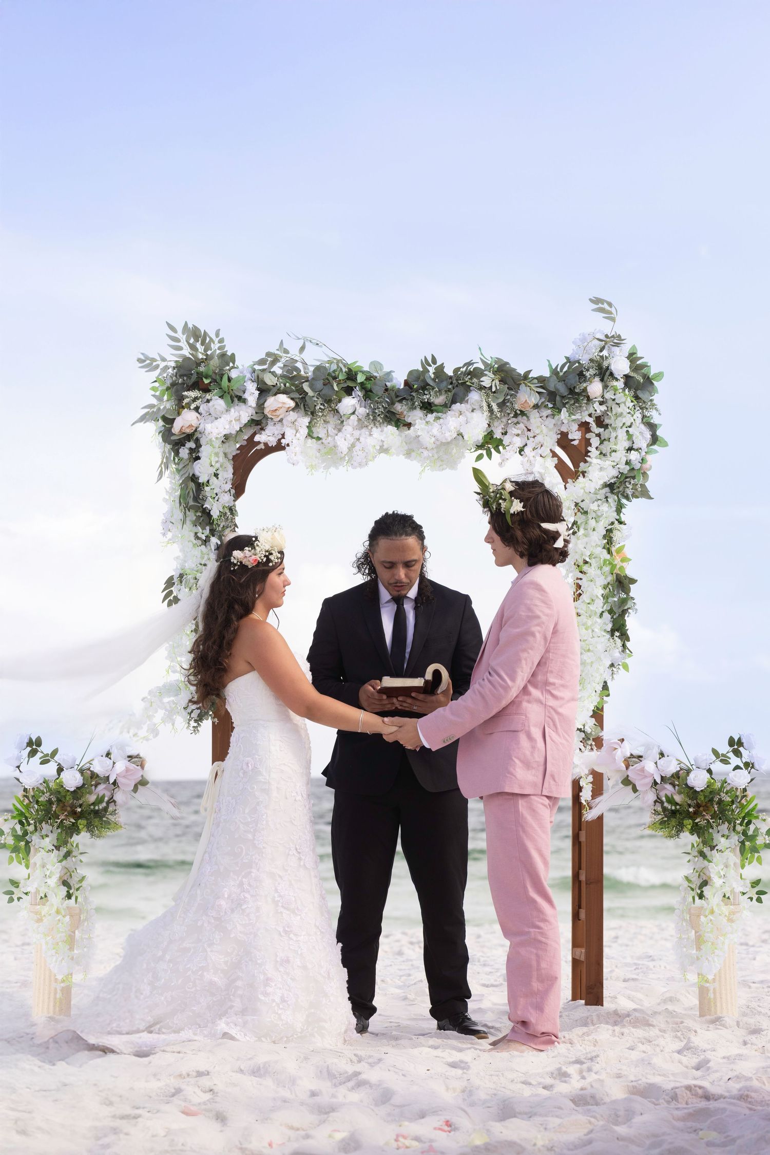 A beachside wedding ceremony takes place under a floral arch decorated with white fabric and greenery on a sandy shore.
