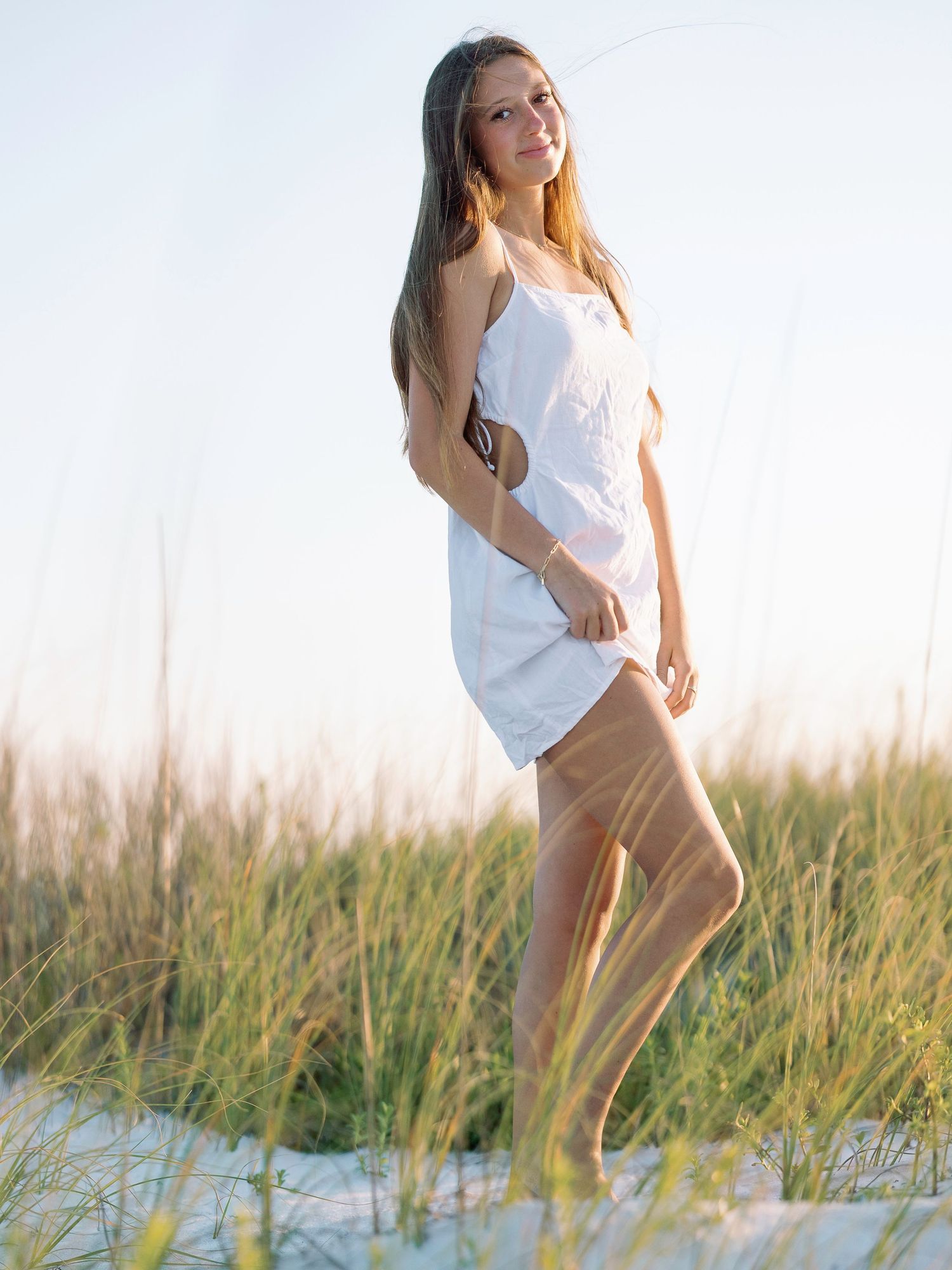 Woman in casual white outfit standing among beach grass during golden hour.
