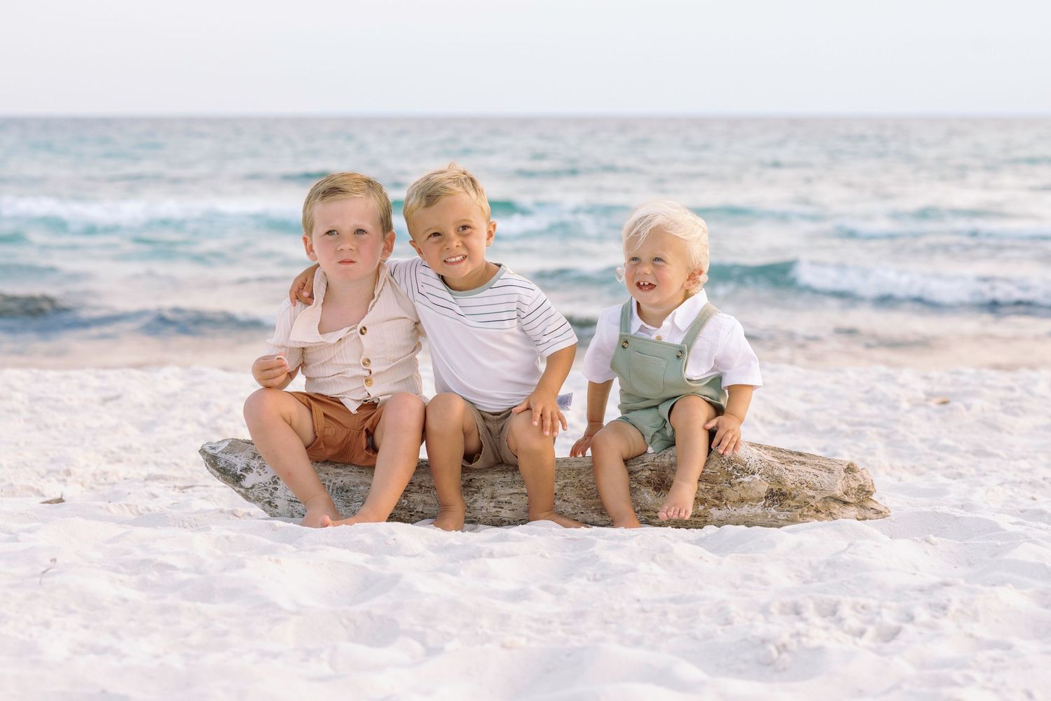 Three children sit together on a rock at the beach with ocean waves behind them.