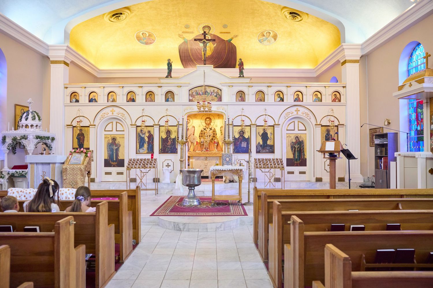 Interior view of an ornate Orthodox church sanctuary with golden dome ceiling, wooden pews and elaborate iconostasis wall.