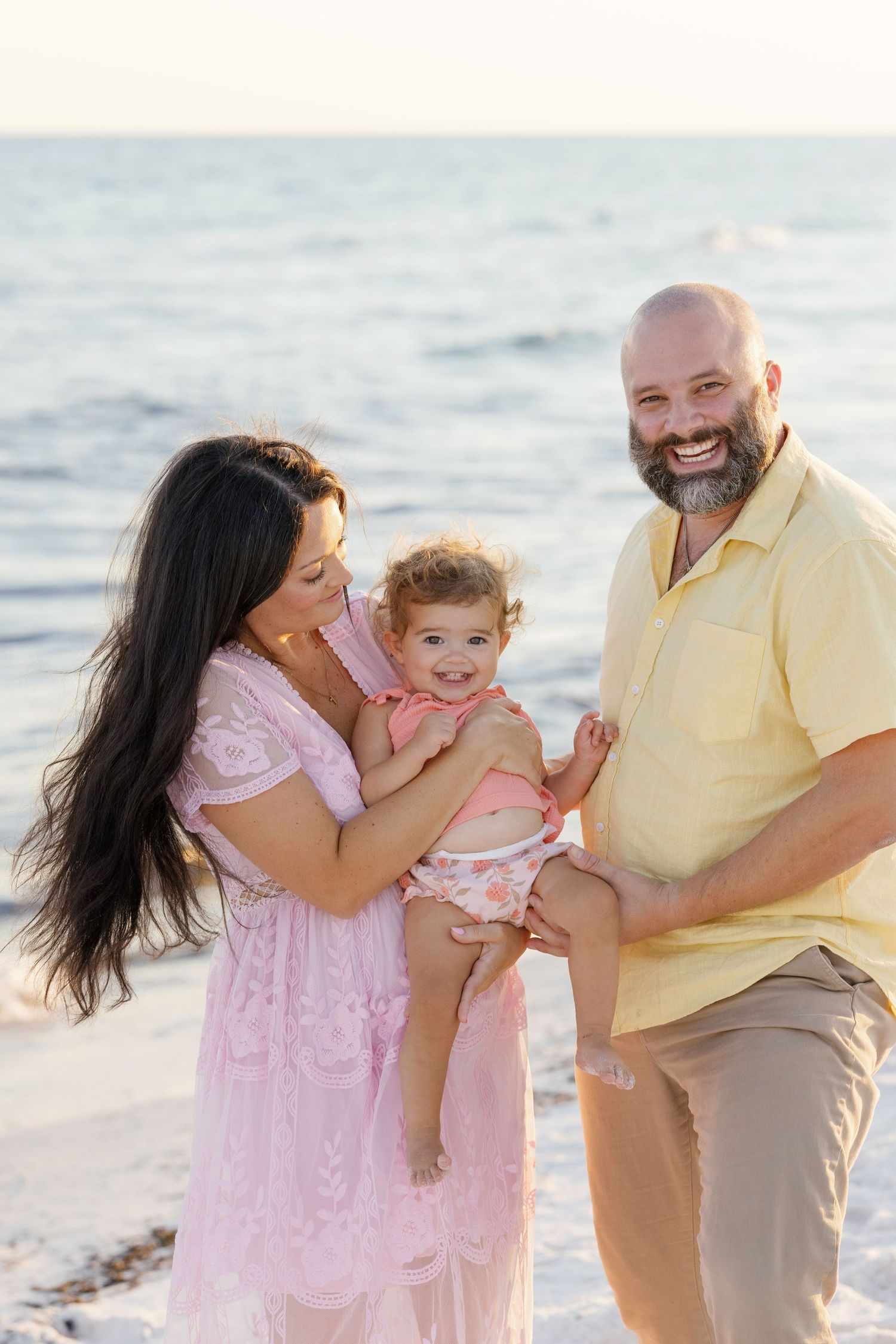 A family wearing pink and yellow outfits shares joyful moments together on a sunny beach at sunset.