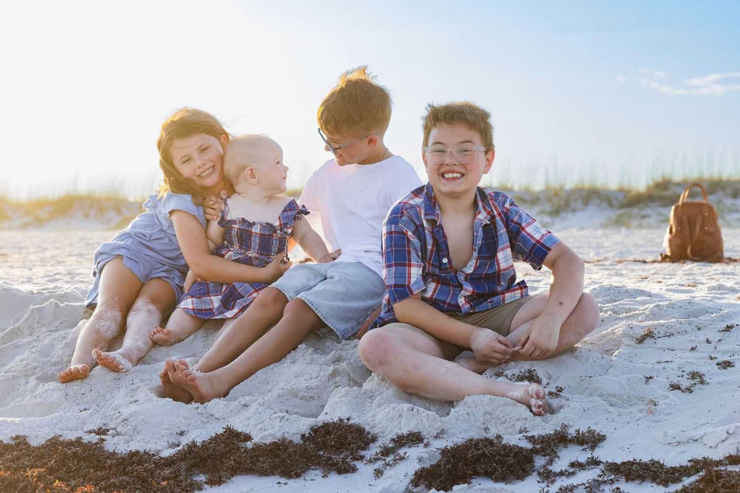 Four young children sit together on white sand at a sunny beach, laughing and playing together.