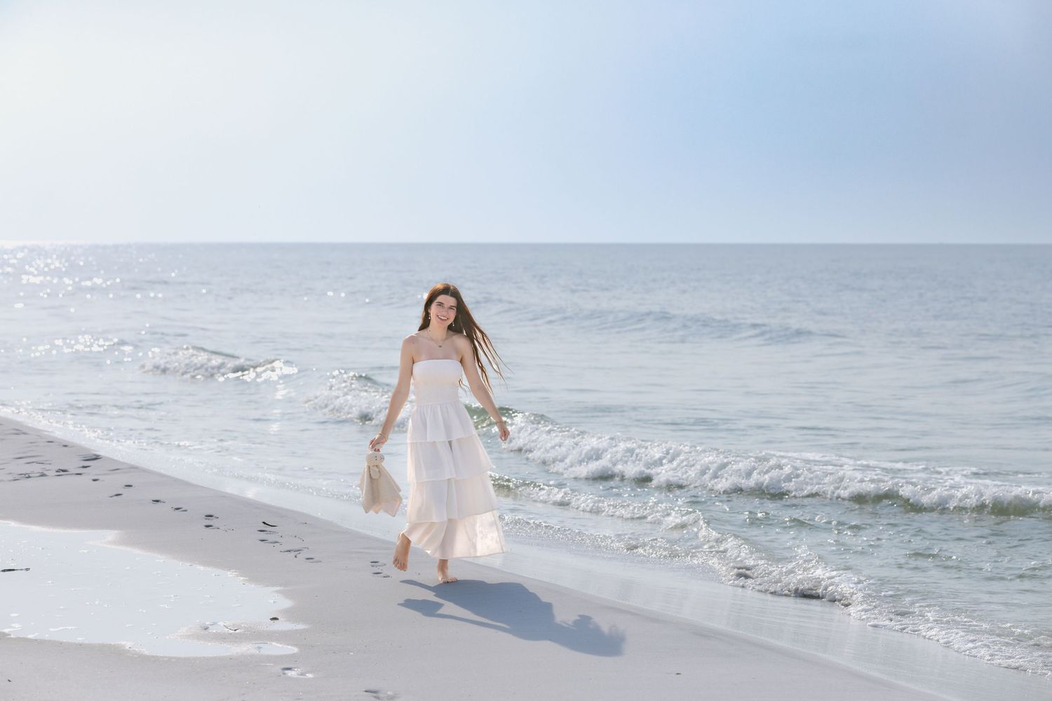 A young woman in a white dress walks along a sunny white sand beach with ocean waves behind her.