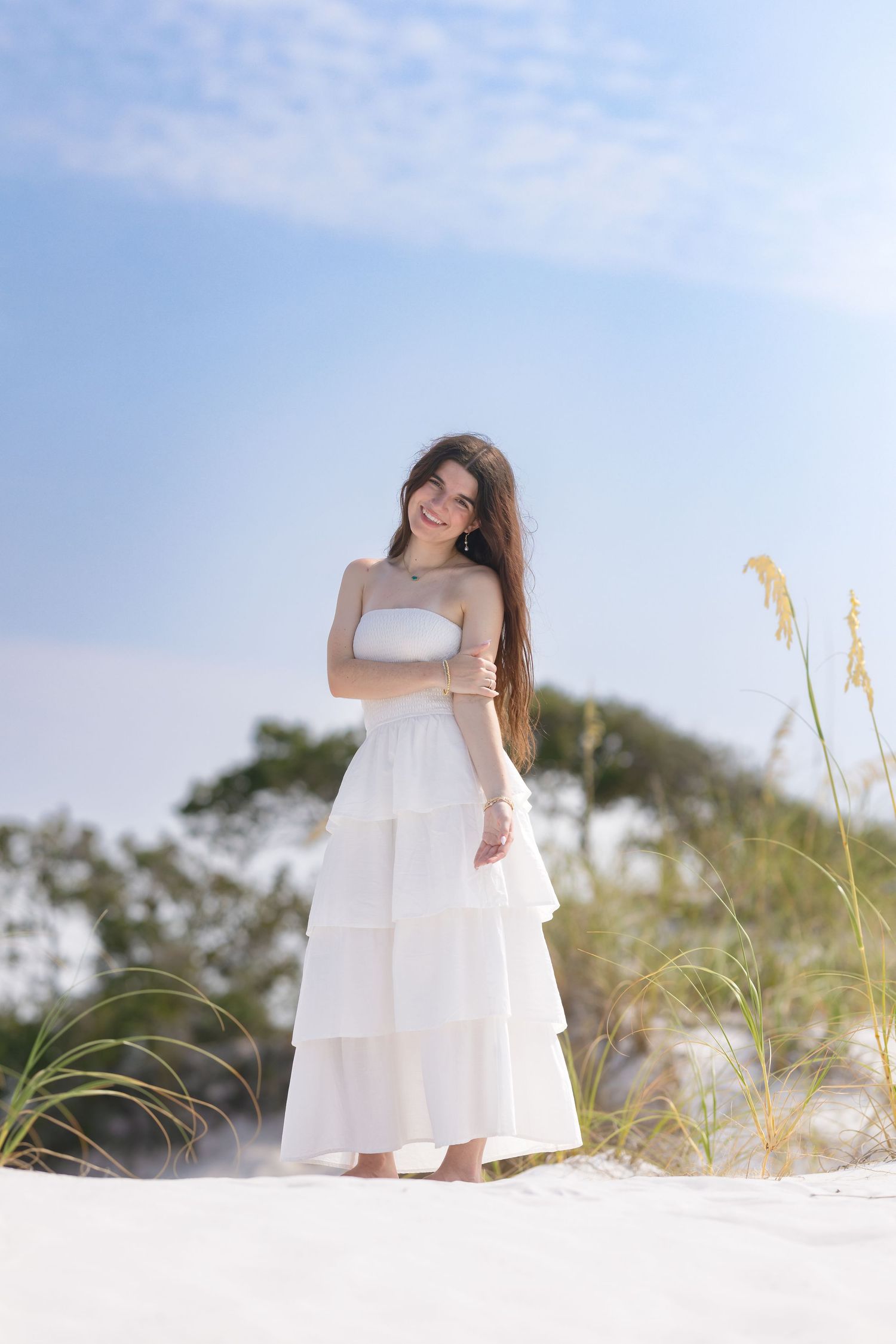 Young woman in white tiered dress standing on white sand dunes with sea oats and blue sky background.