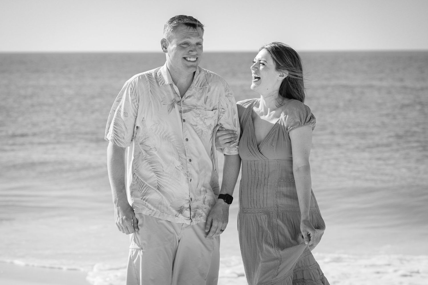 Black and white photo of a smiling couple holding hands and walking on a beach near the ocean.