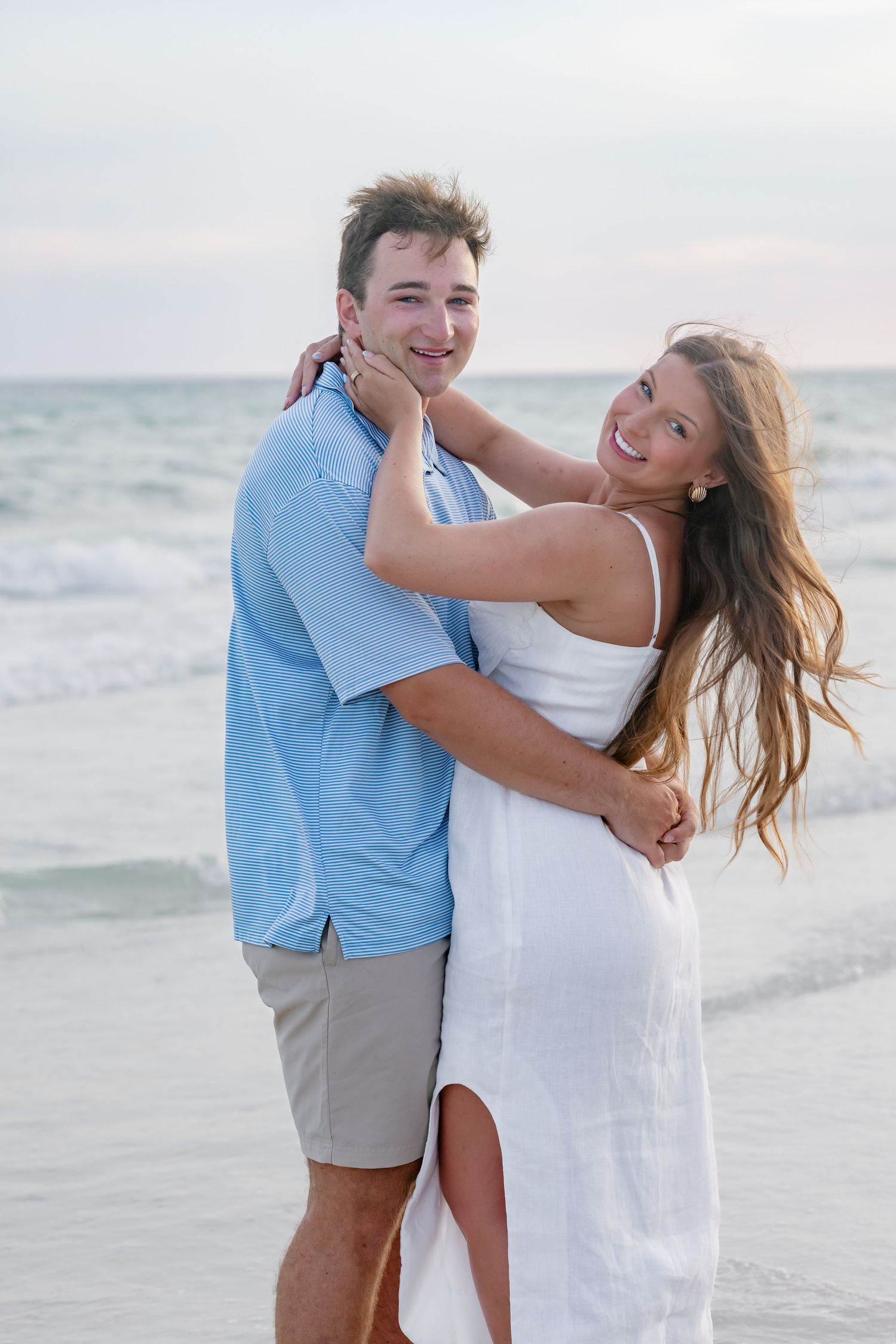A happy couple embraces on a beach, woman in white dress and man in blue shirt, with ocean waves in the background.