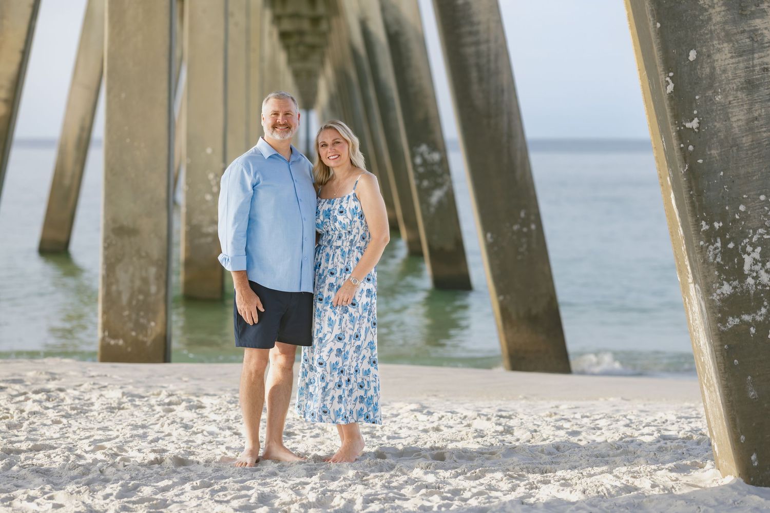 Couple smiling together on sandy beach under wooden pier with turquoise water in background.