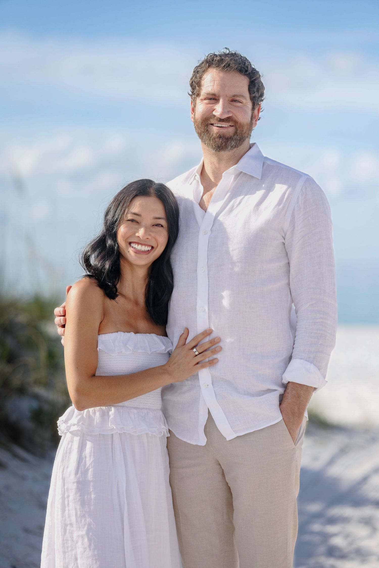 A smiling couple in white outfits poses together on a sunny beach, with blue skies and sand dunes in the background.