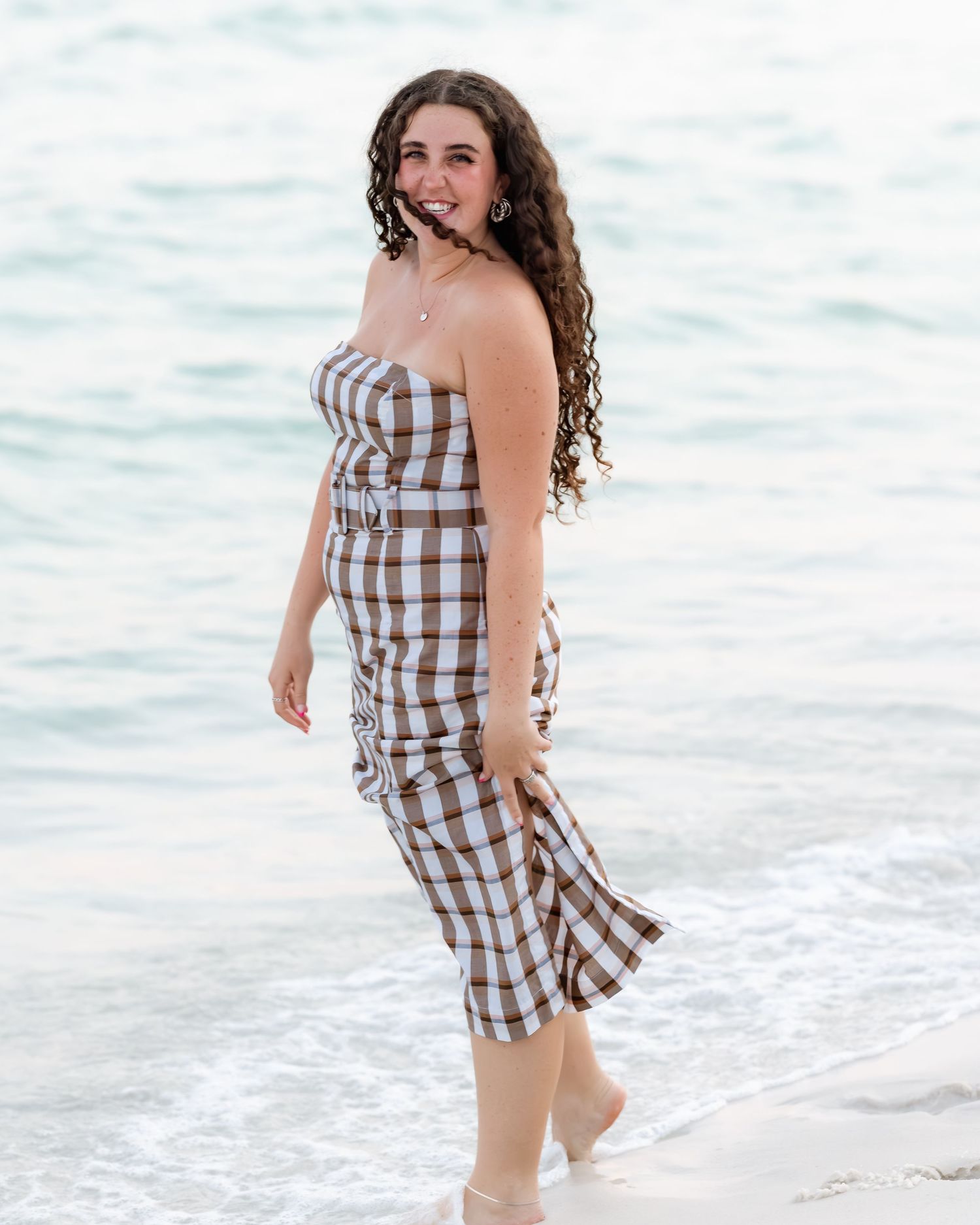 Young woman in brown plaid strapless dress poses confidently at beach shoreline with ocean waves behind her.