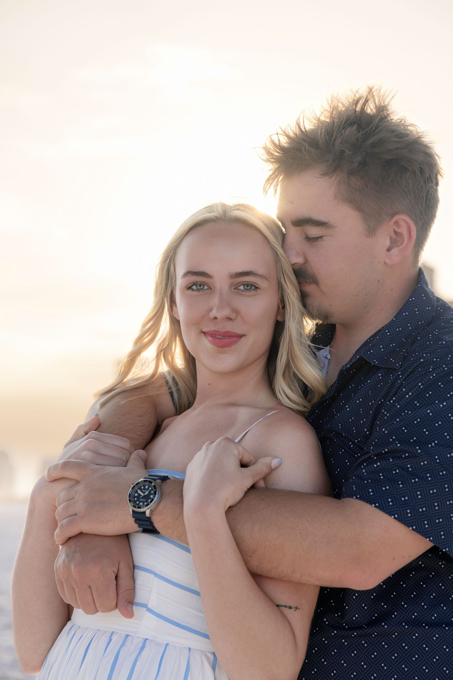 A couple embraces at sunset on a beach, the man kissing the woman's cheek as she smiles at the camera.