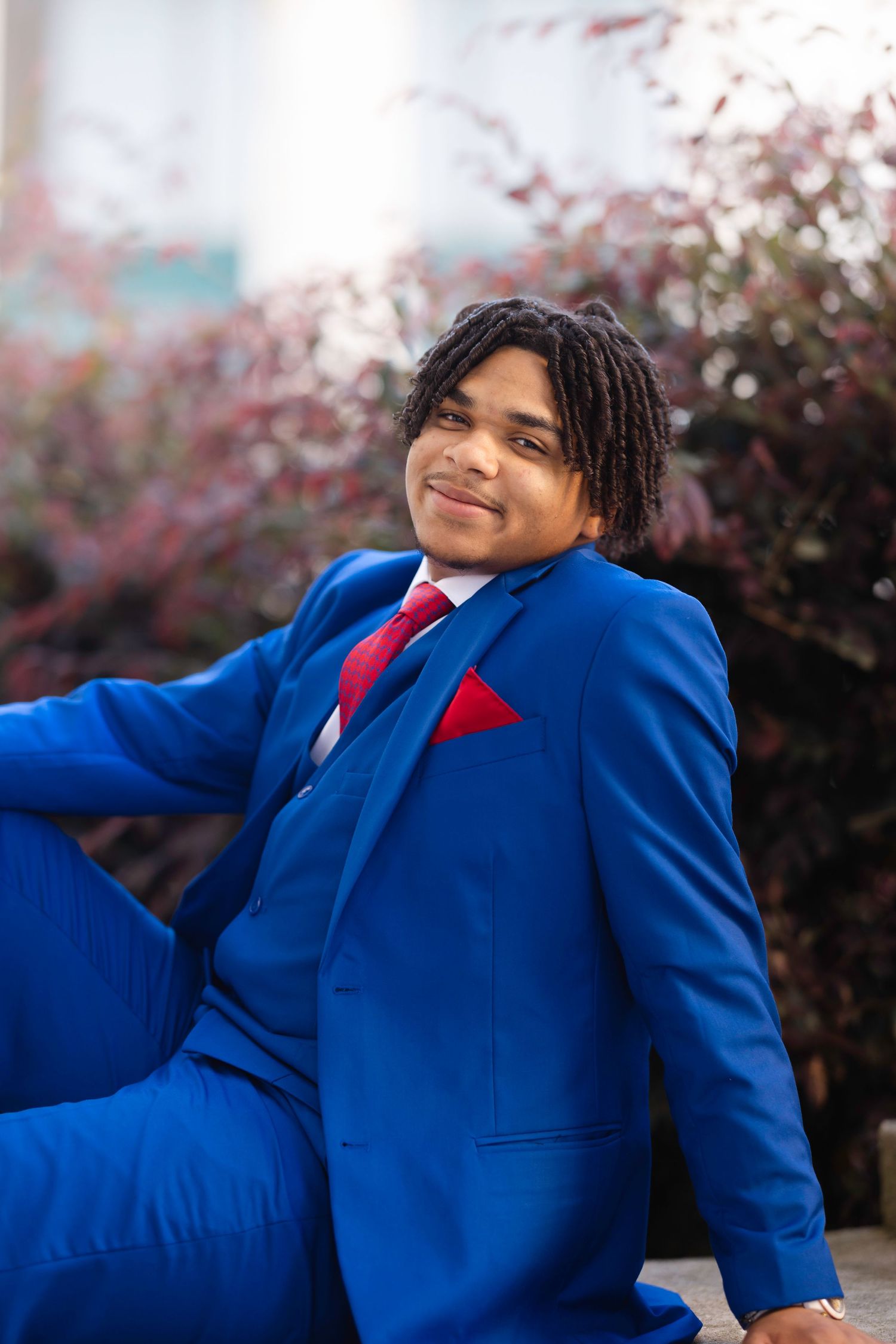 Young man in blue suit with red tie and pocket square smiling outdoors near flowering shrubs.