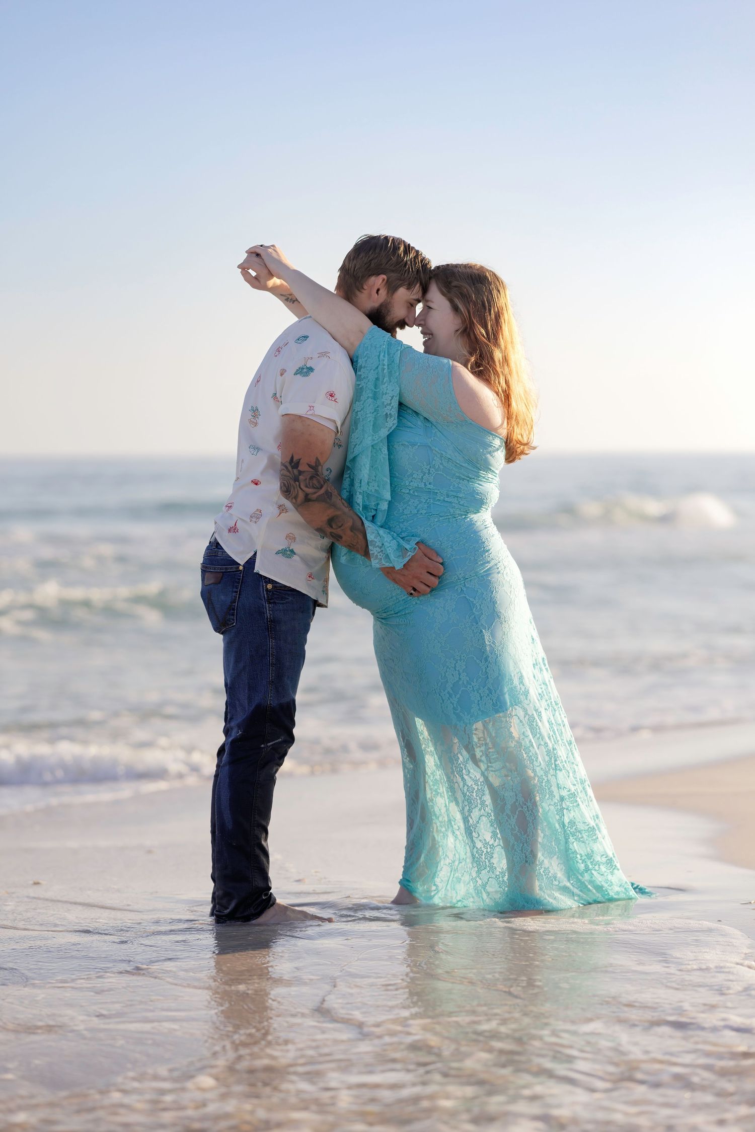 Expecting couple in teal dress and floral shirt dance romantically at the ocean's edge on a sunny beach.