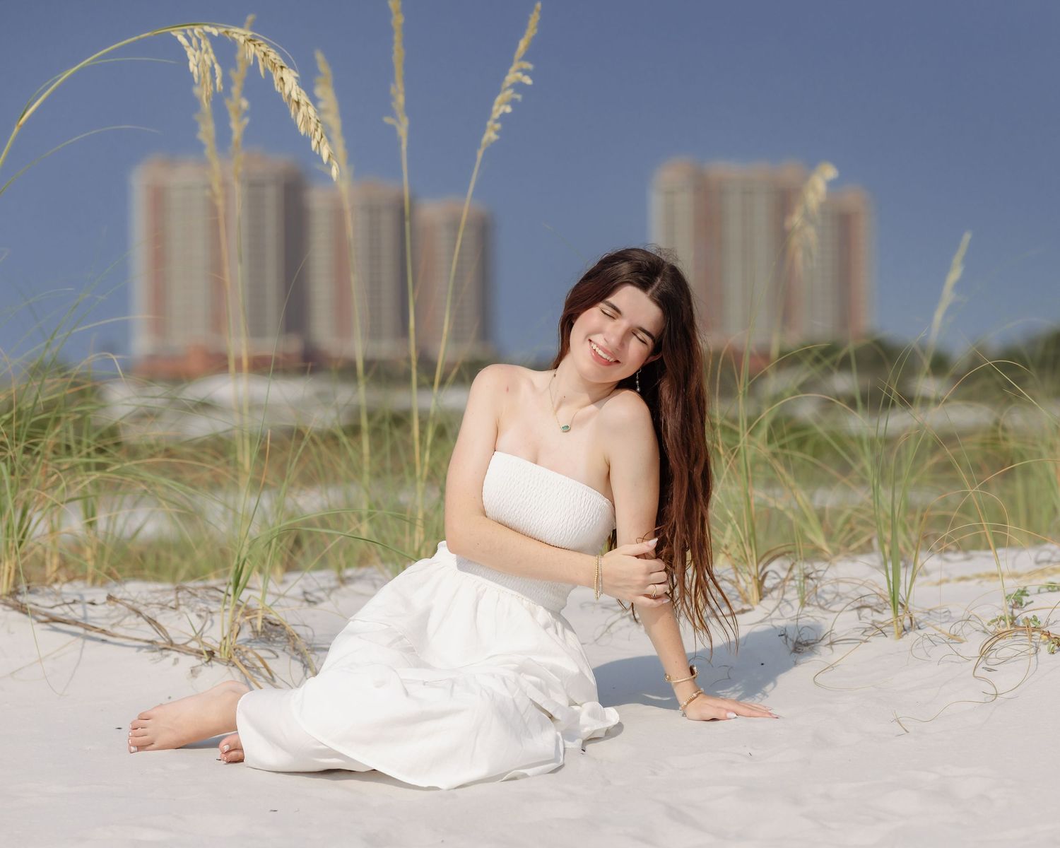 Young woman in white strapless dress sitting on white sand beach dunes with tall buildings and sea oats in background.