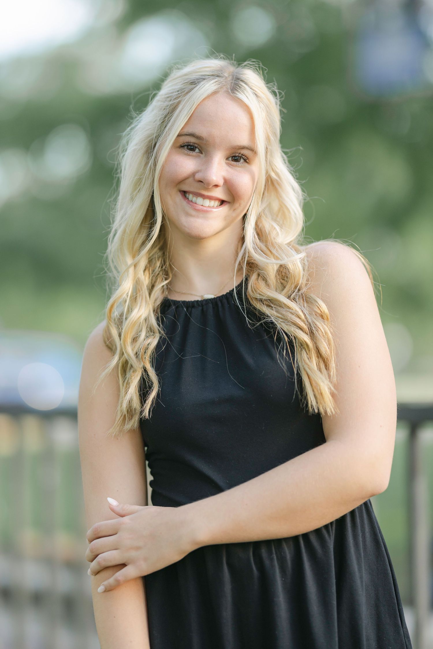 Smiling young blonde woman in a black sleeveless dress posing outdoors with a blurred green background.