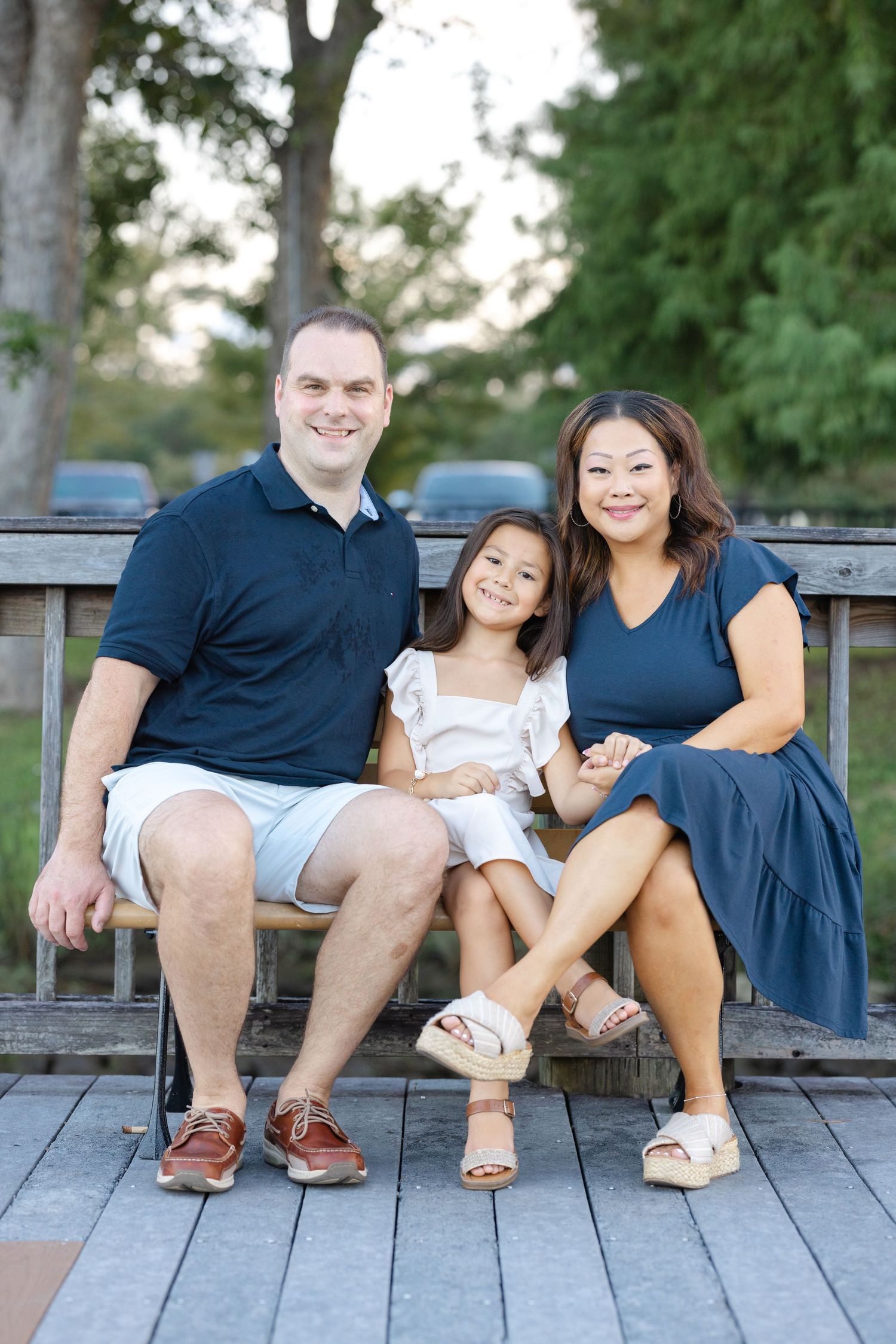 A smiling family of three poses on a wooden bench outdoors, with parents flanking their young daughter in a park setting.