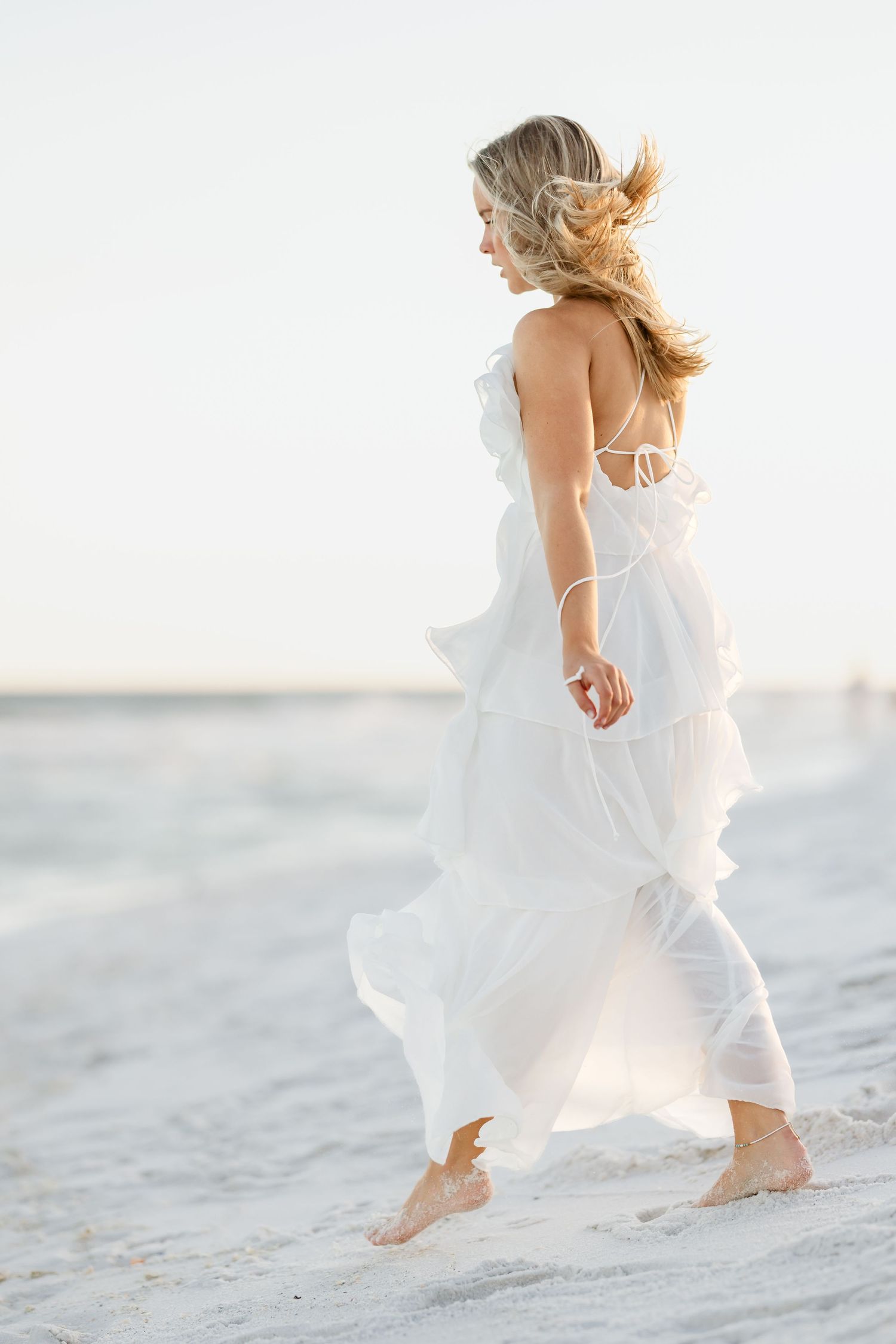 Blonde woman in flowing white ruffle dress walking barefoot on sandy beach with wind in her hair.