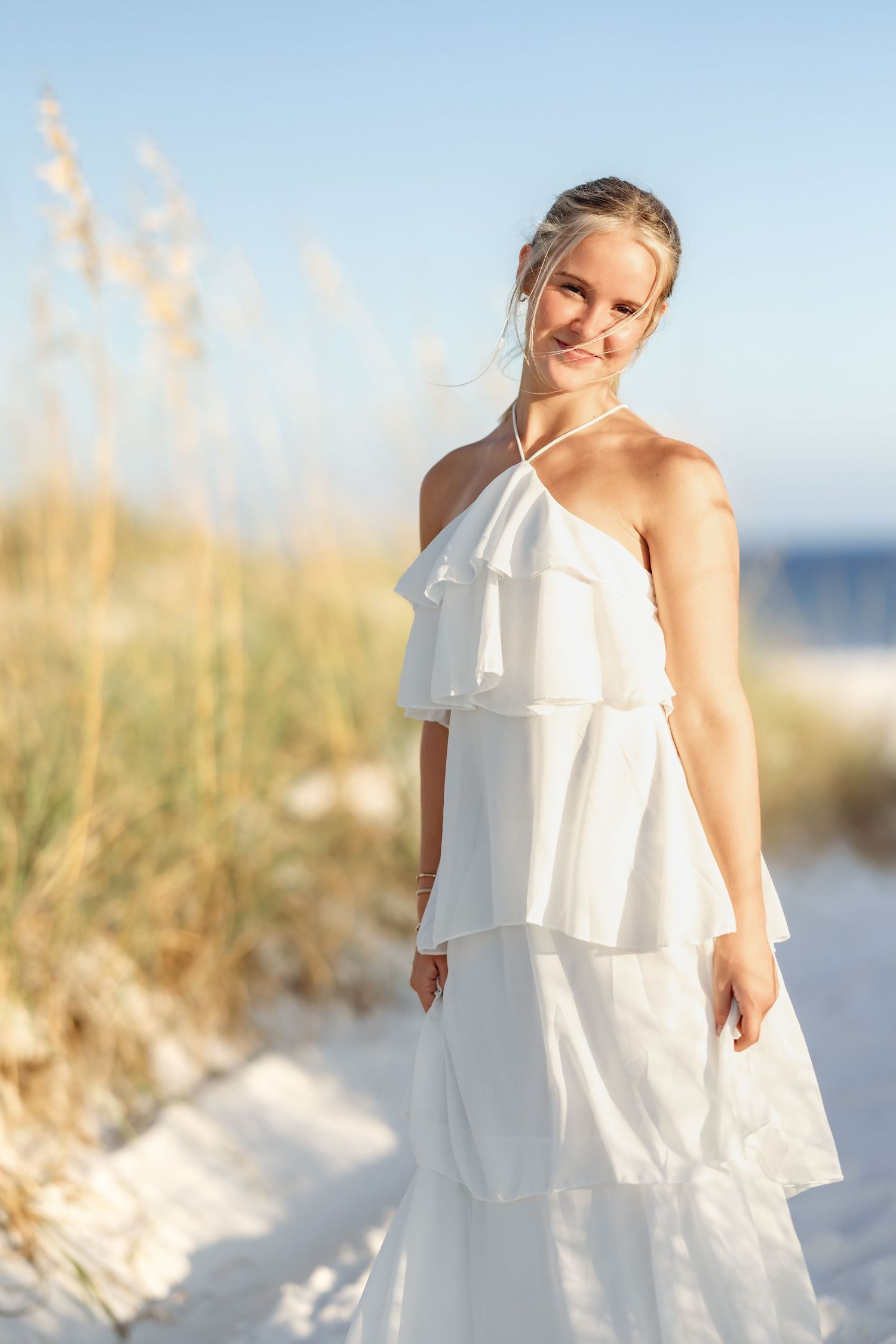 Smiling blonde woman in white ruffled halter maxi dress standing on sandy beach dunes with sea oats.