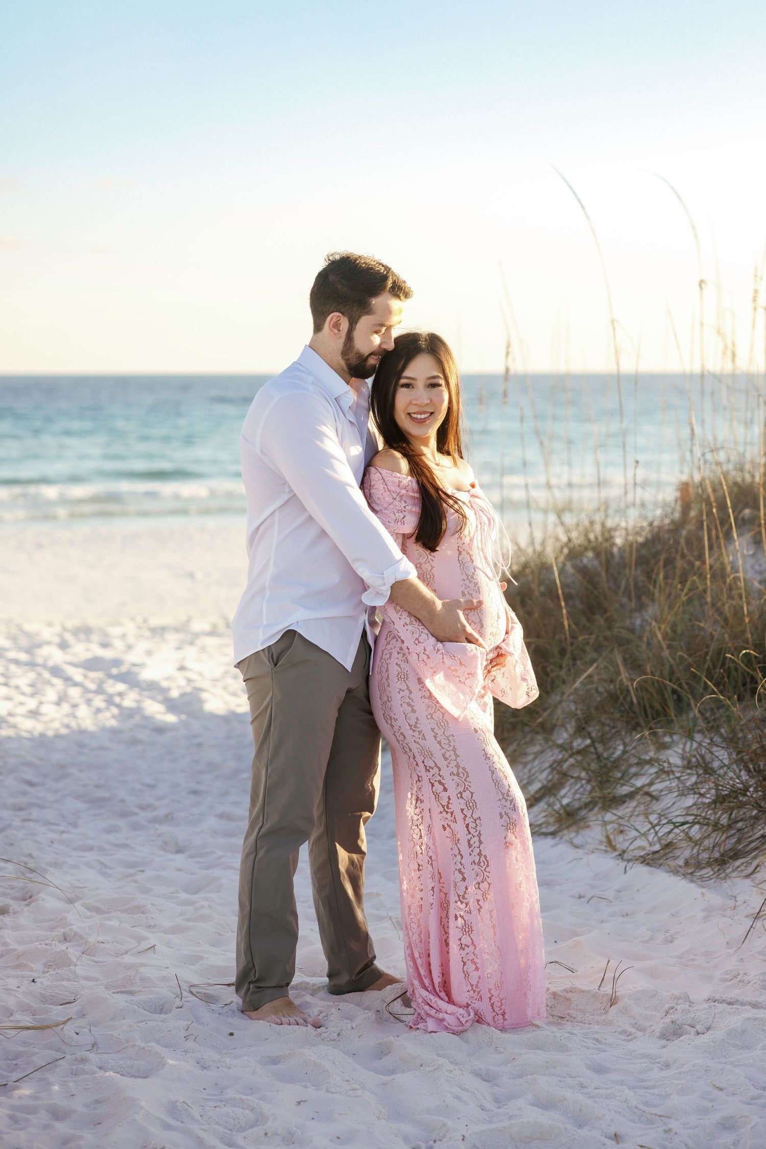 A couple poses together on a white sandy beach at sunset with sea oats and ocean waves in the background.