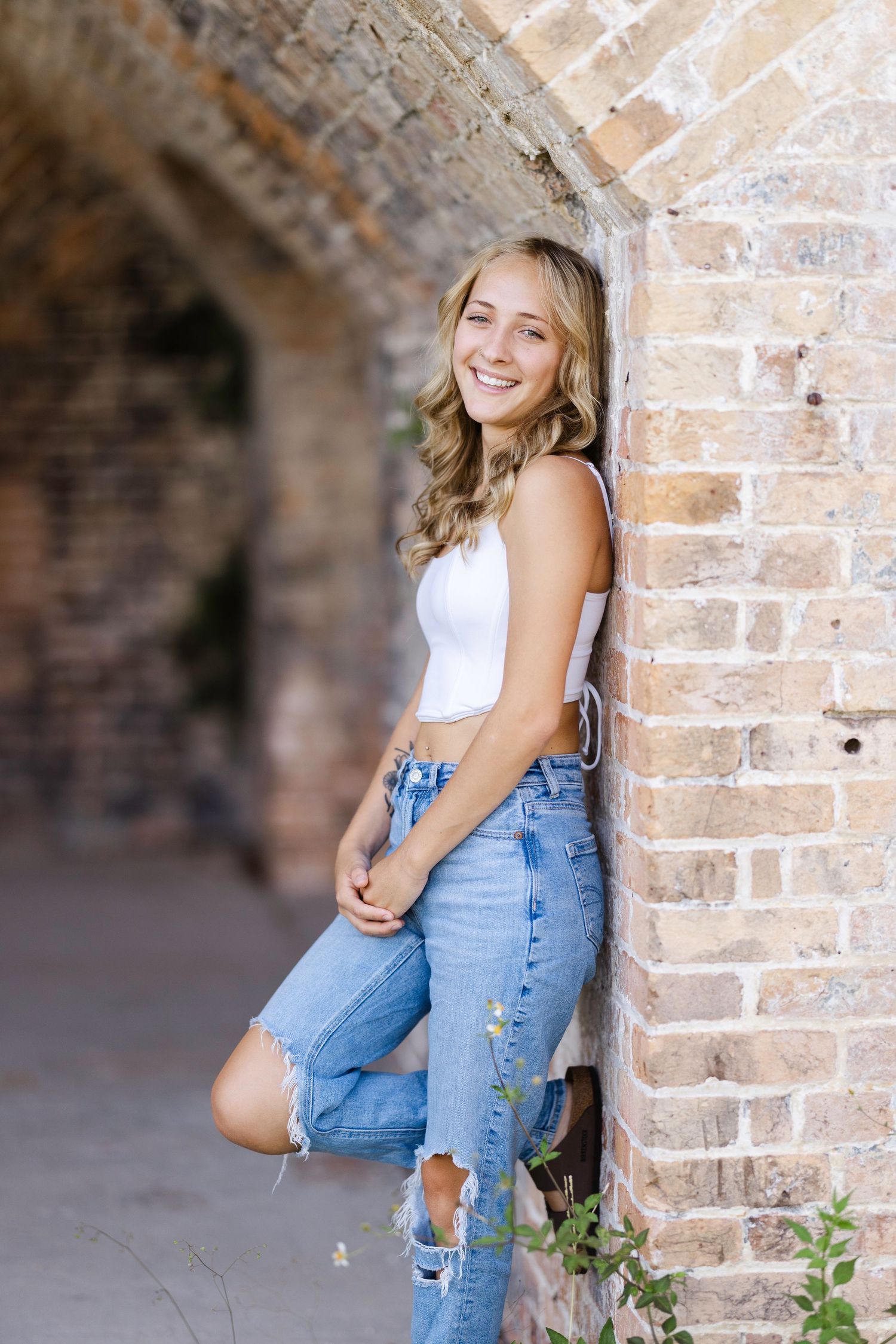 A person in a white tank top and ripped jeans poses casually against a rustic brick archway.