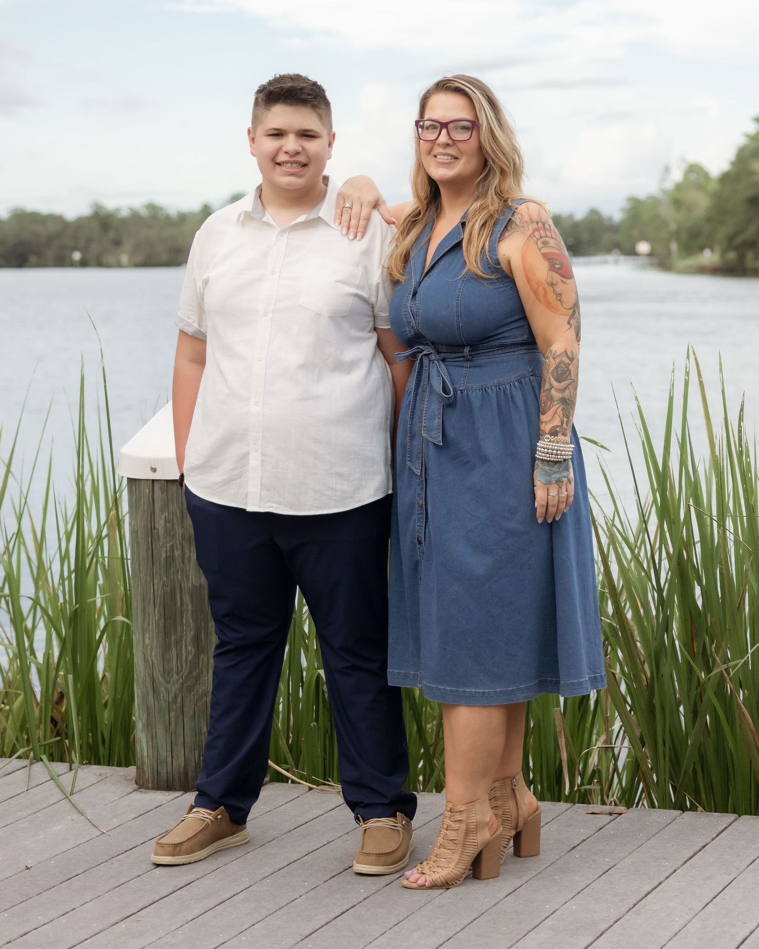 A young man and tattooed woman smiling together on a wooden dock by a lake for a portrait.