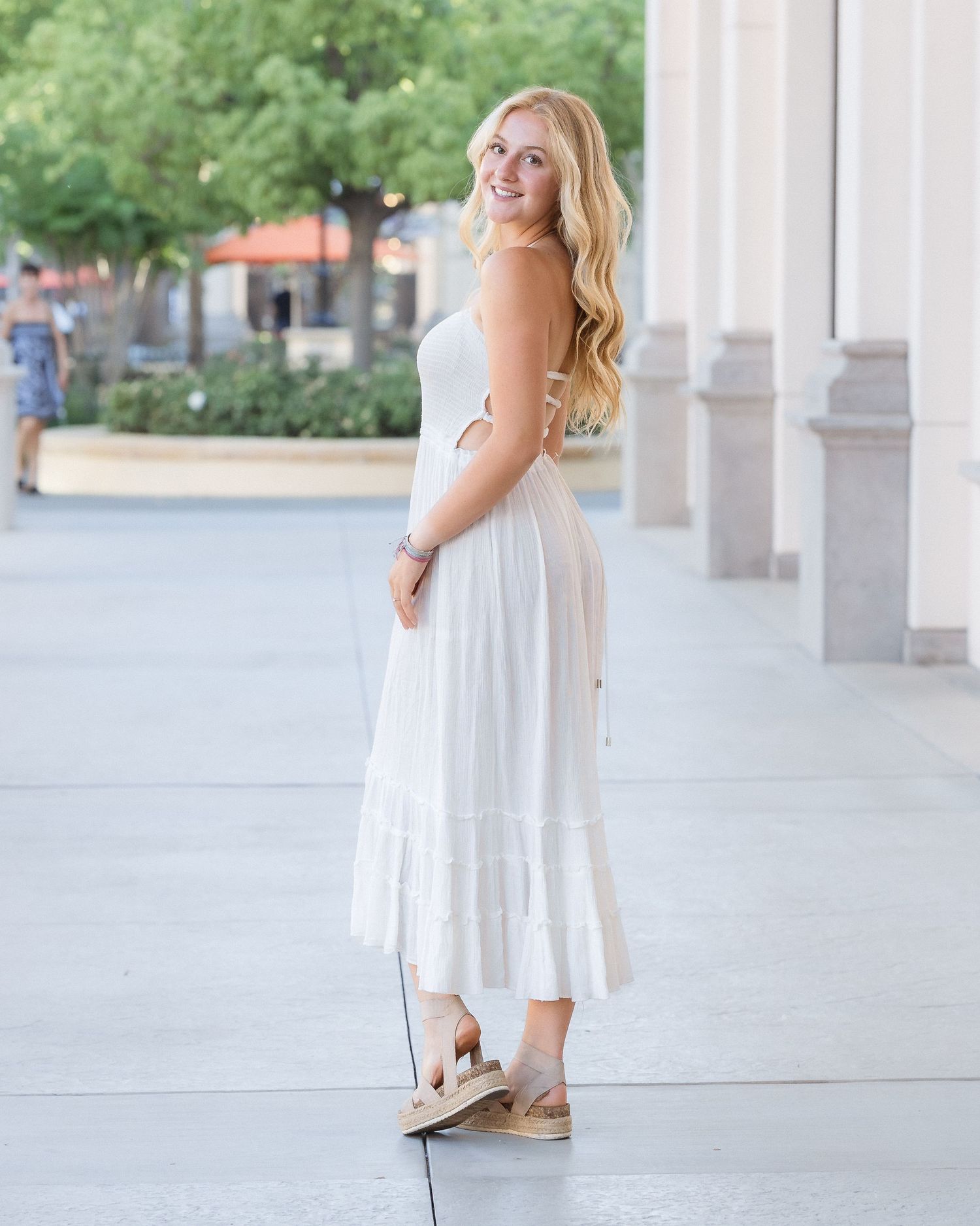 Young blonde woman smiling in a white tiered maxi dress and espadrille sandals, posing outdoors near columns.