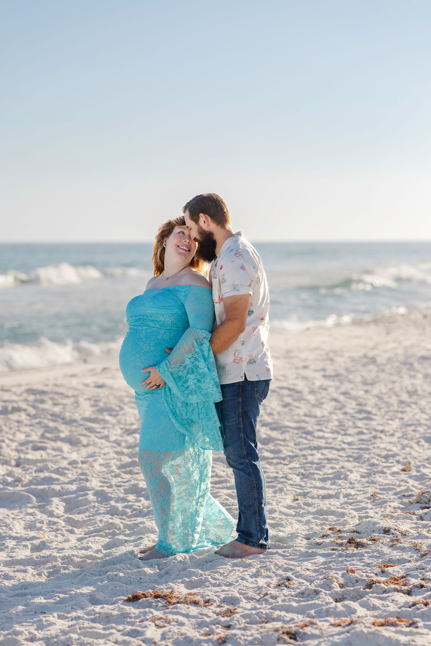 Pregnant couple embracing on a white sand beach, woman in teal maternity gown, man kissing her head near ocean waves.