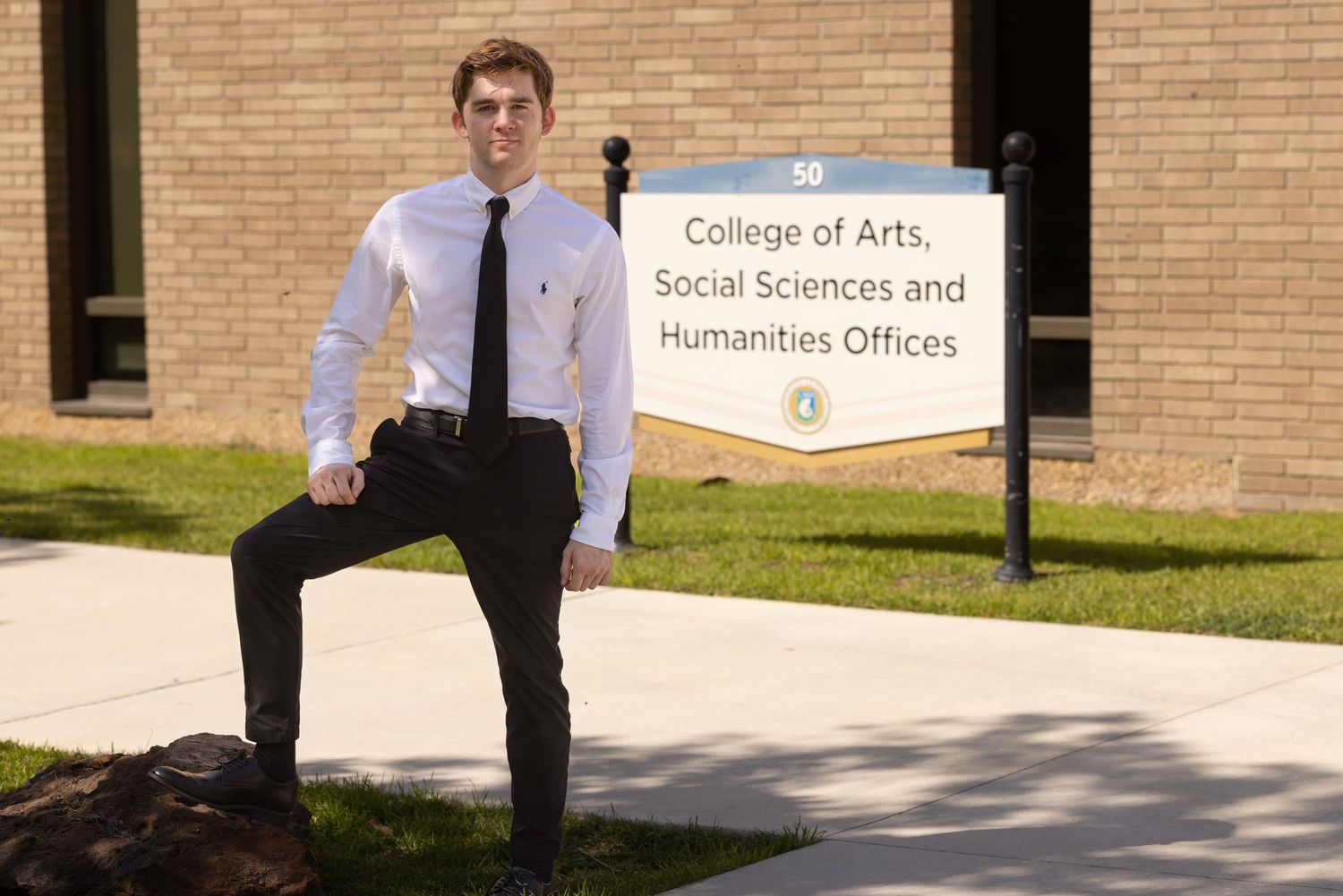 Young man in white shirt and tie posing outside College of Arts, Social Sciences and Humanities Offices sign.