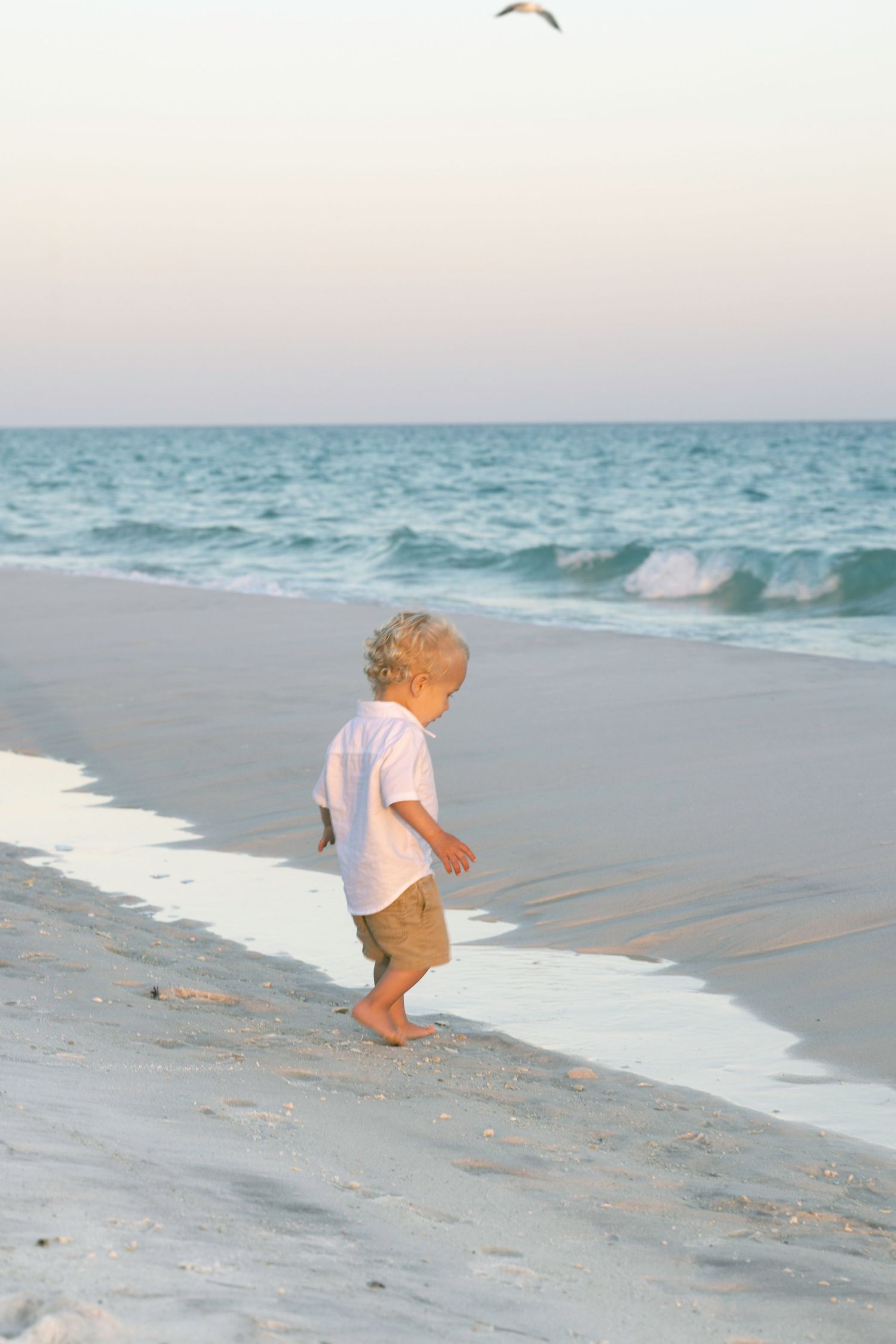 A barefoot toddler boy in white shirt and khaki shorts walks along a sandy beach at sunset.