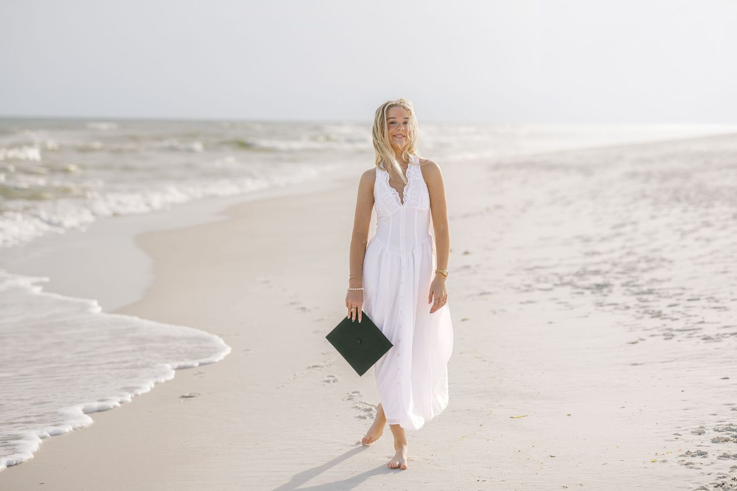 Woman in white maxi dress walking barefoot on sandy beach holding dark clutch purse near ocean waves.