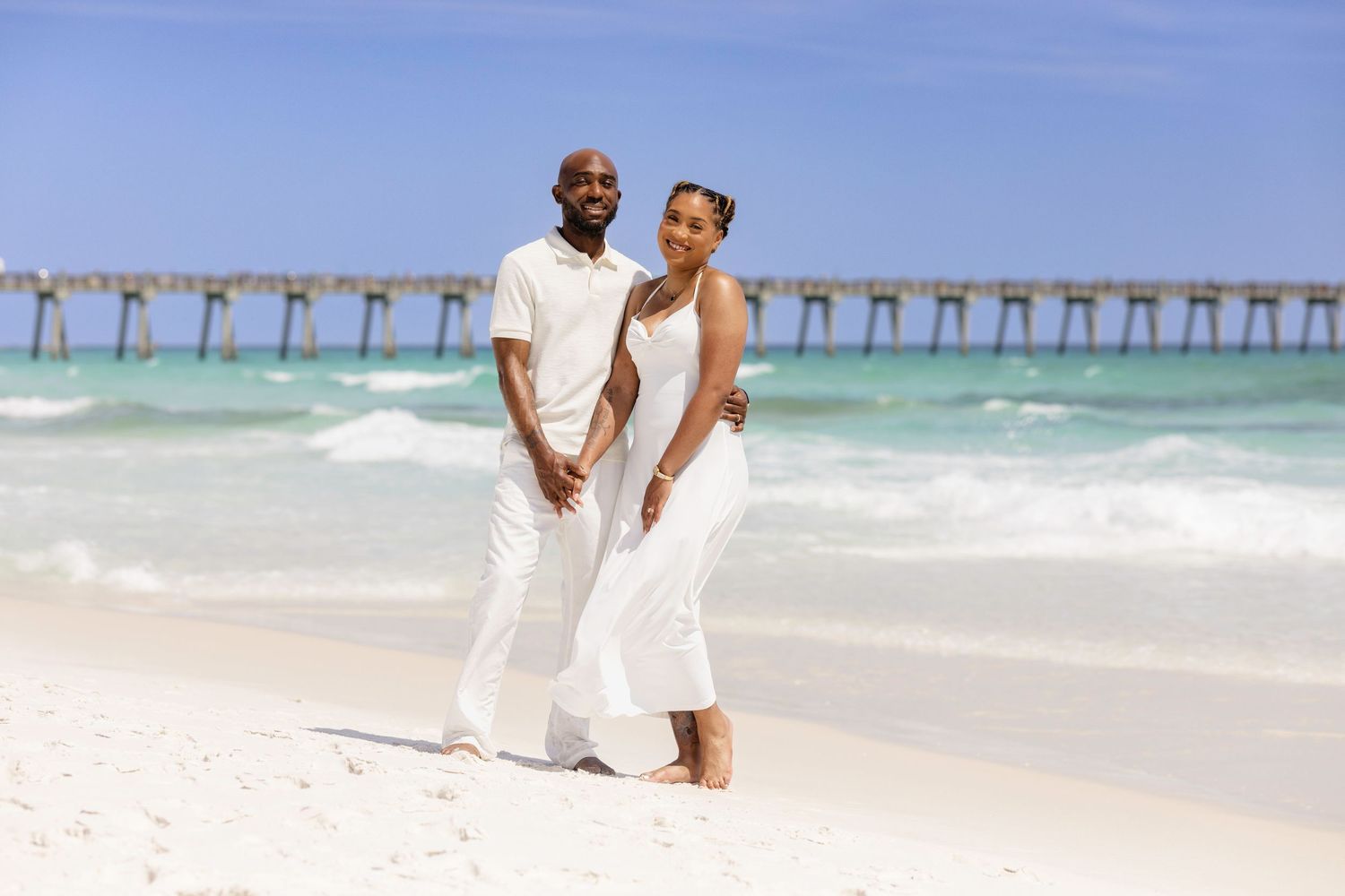 A smiling couple dressed in white holds hands on a sunny white sand beach with turquoise water behind them.
