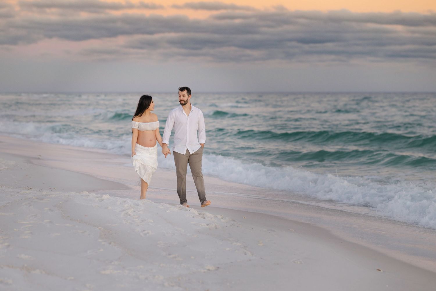 Couple holding hands on a white sandy beach at sunset with turquoise ocean waves in the background.