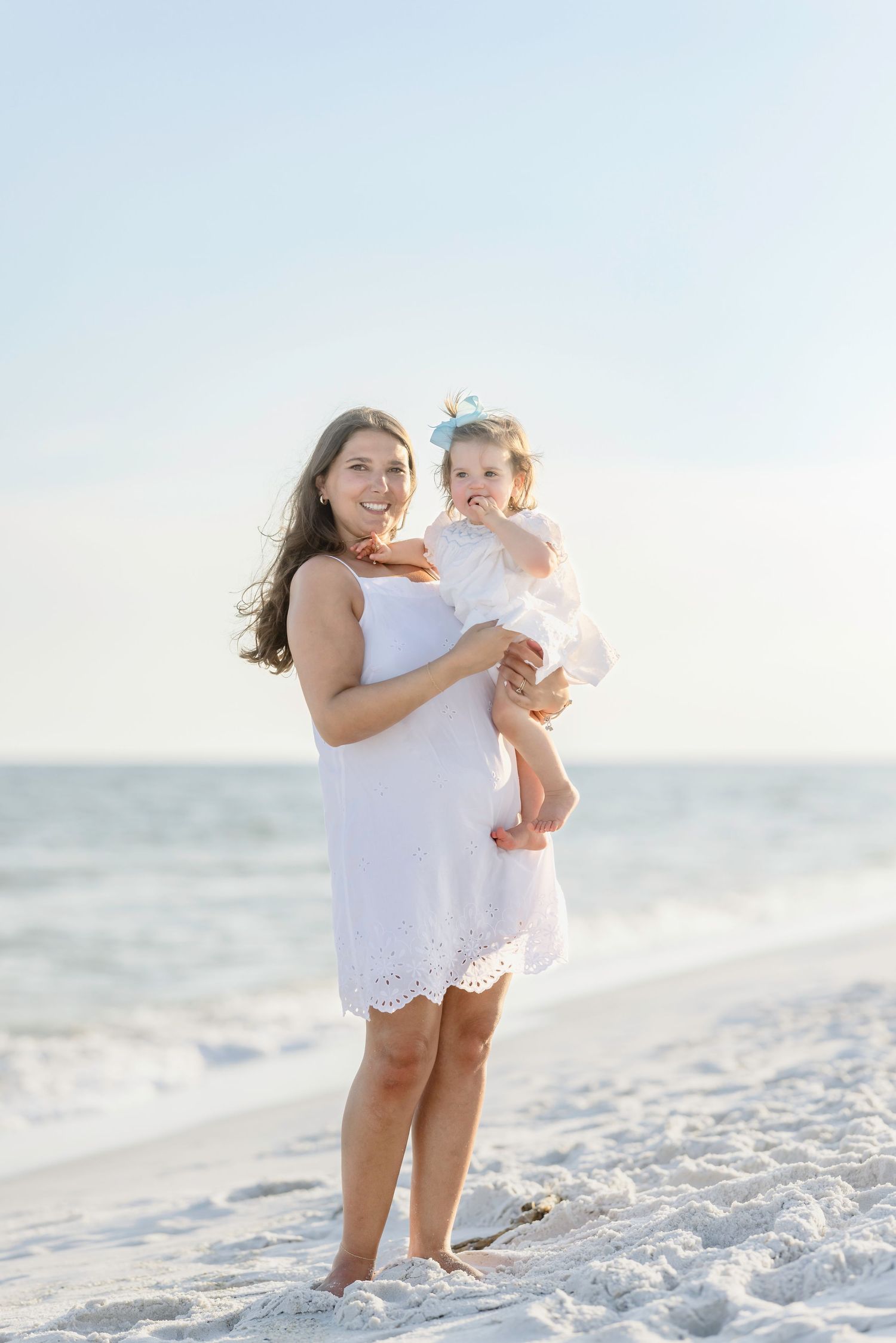 Mother in white dress holds baby girl in white outfit on a sunny beach.