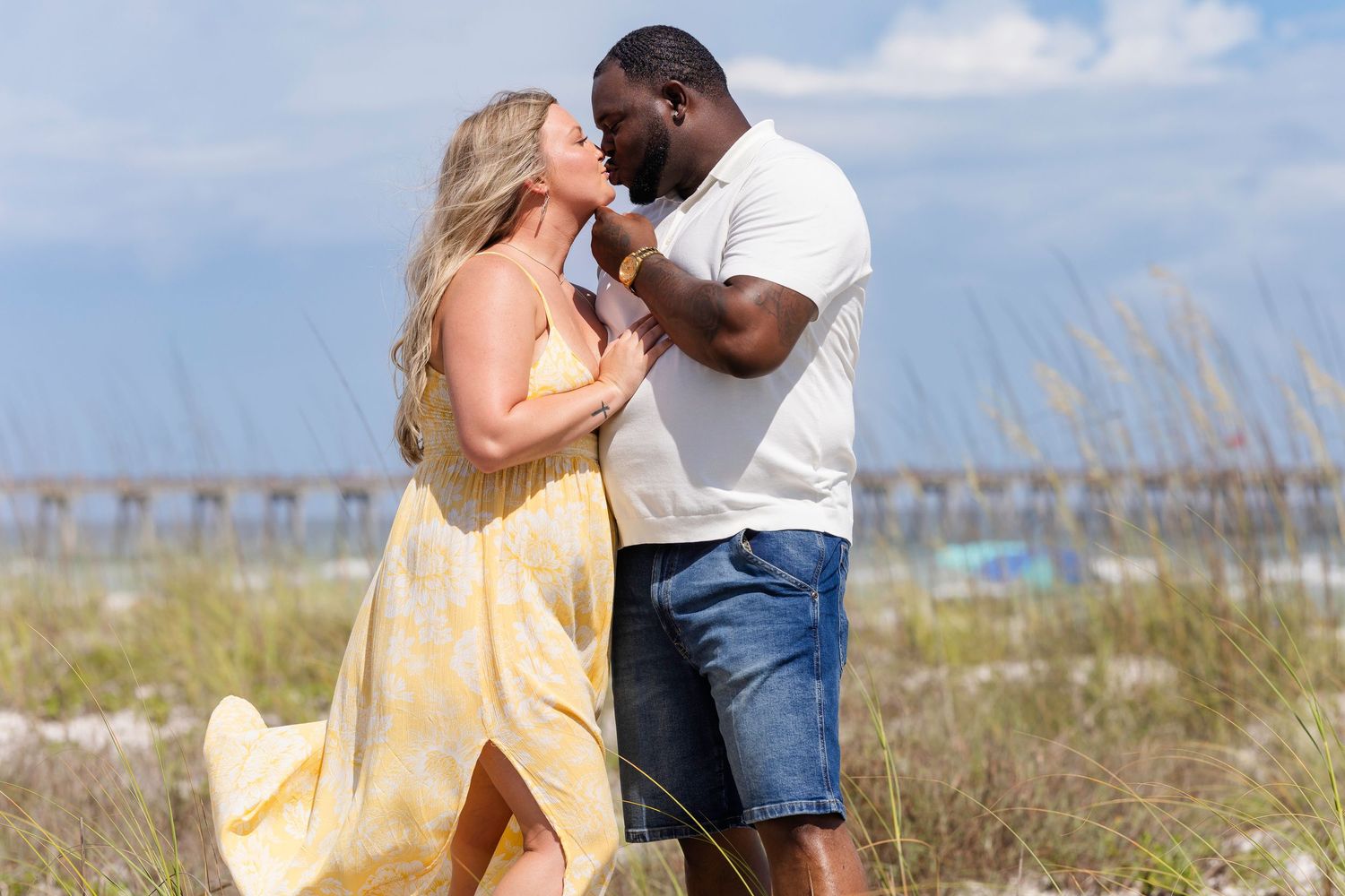 Romantic interracial couple sharing a kiss on a breezy beach with a pier in the background.