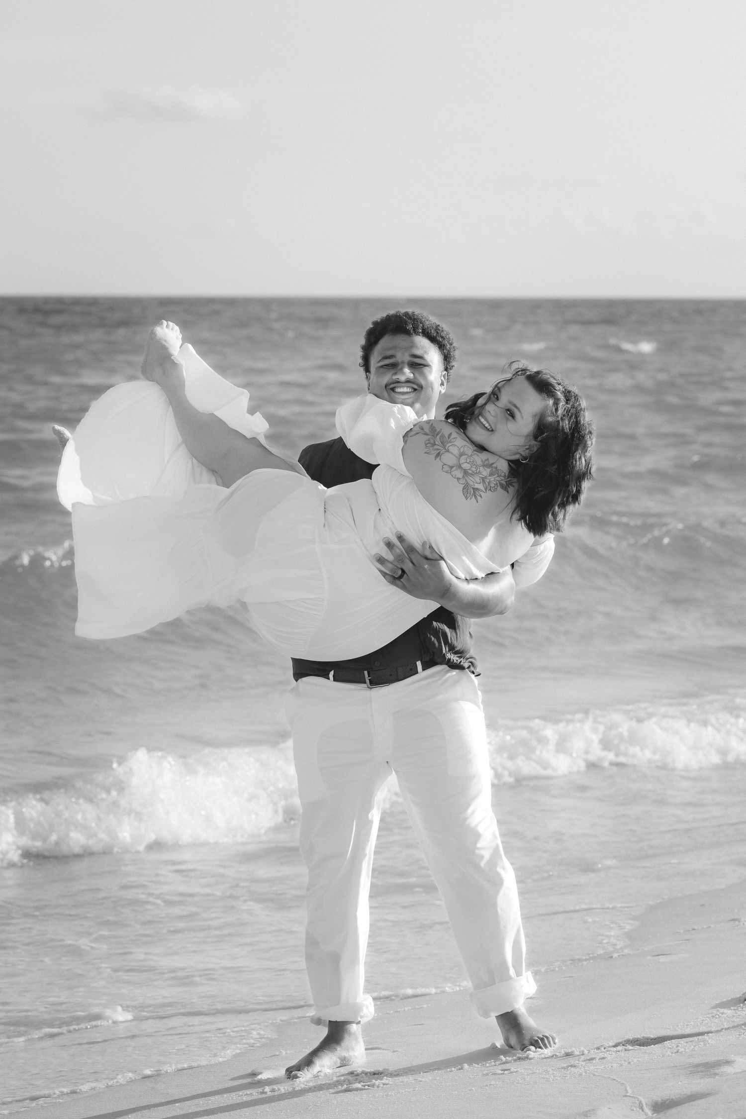 Black and white photo of a groom carrying his bride in a wedding dress on a beach with ocean waves.