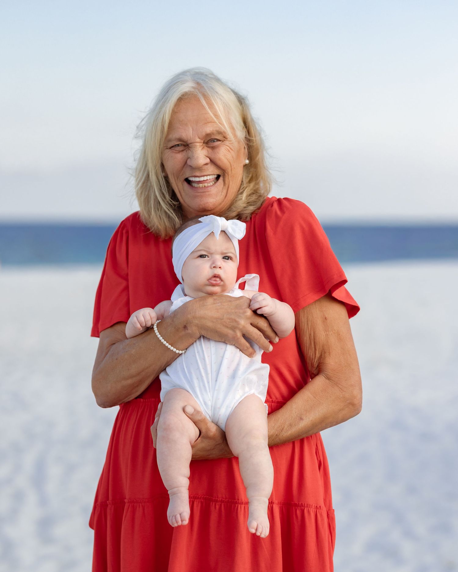 Smiling grandmother in red dress holding adorable baby in white outfit on a sunny beach.
