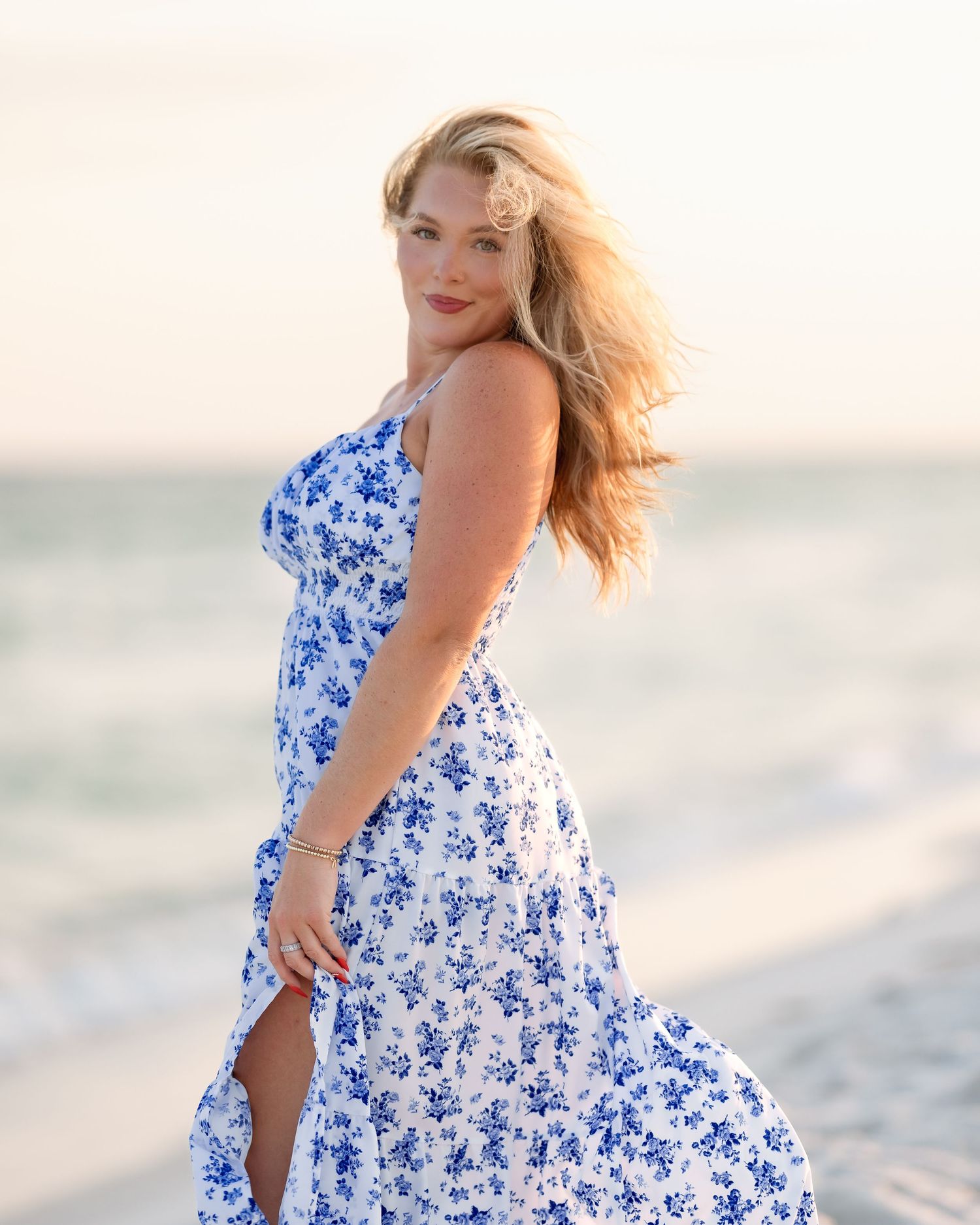 A blonde woman in a blue floral maxi dress poses on a sunny beach.