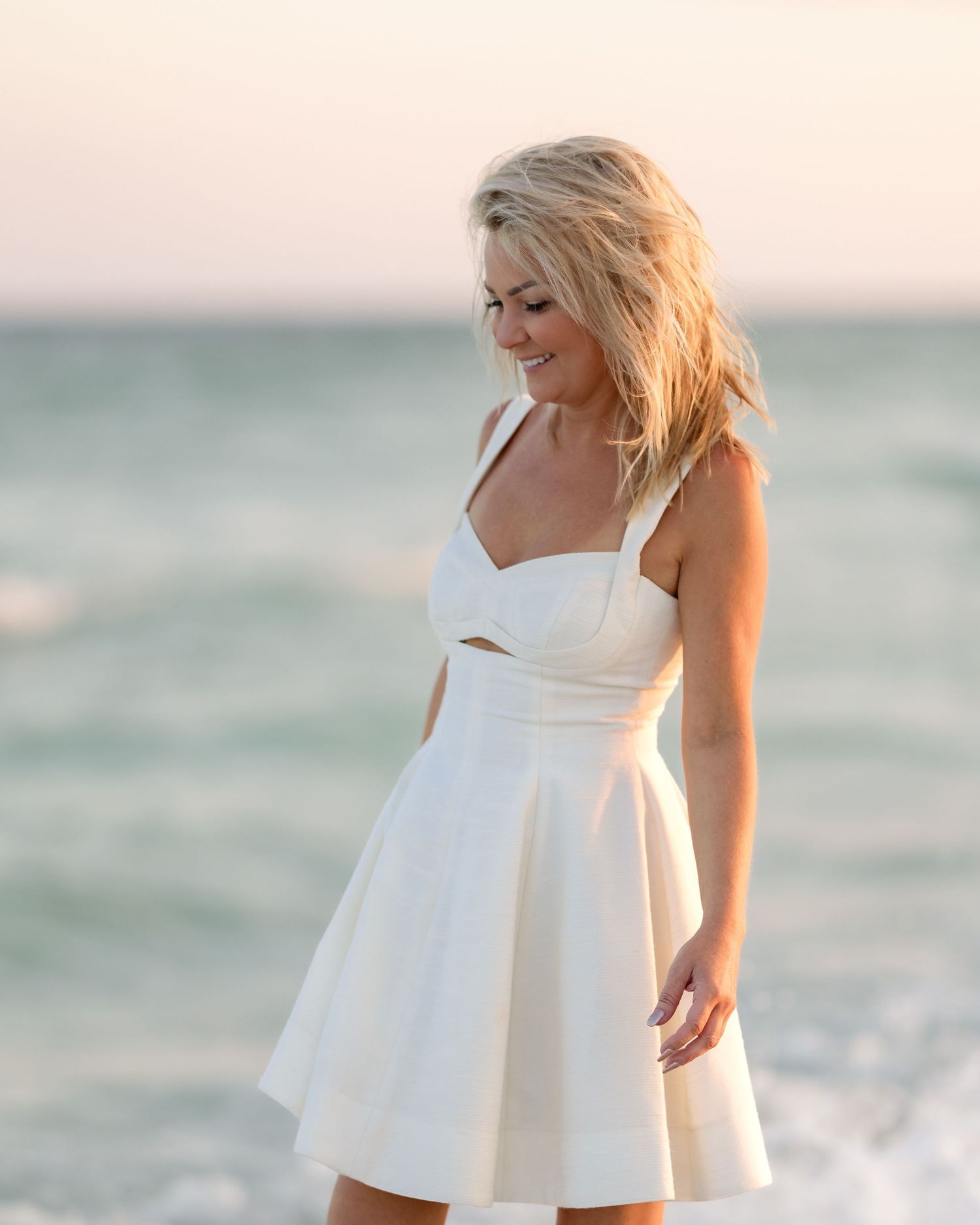 Blonde woman in white sundress smiling at beach with ocean waves behind her.