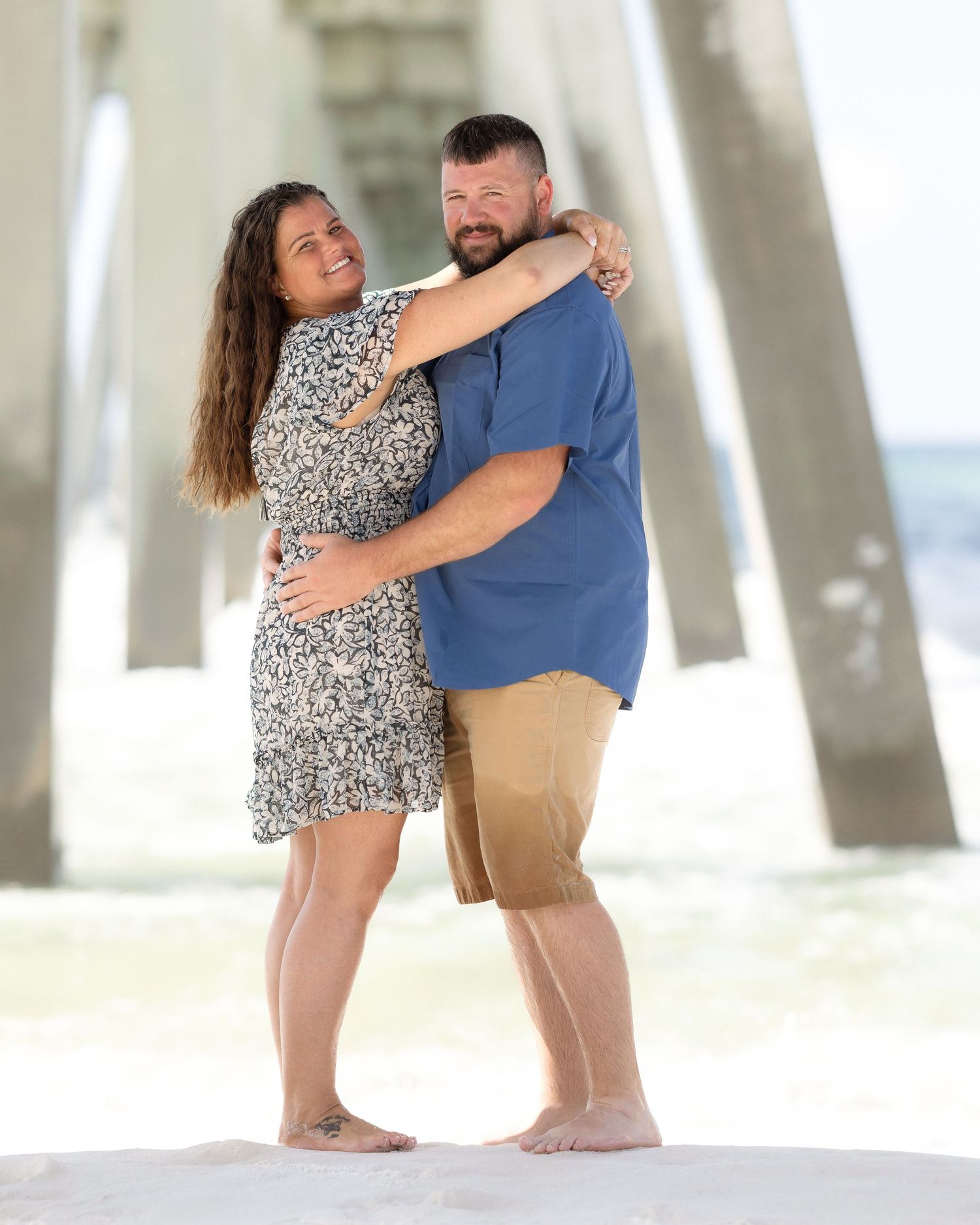 Couple embracing and smiling barefoot on sandy beach under pier, woman in floral dress, man in blue shirt.