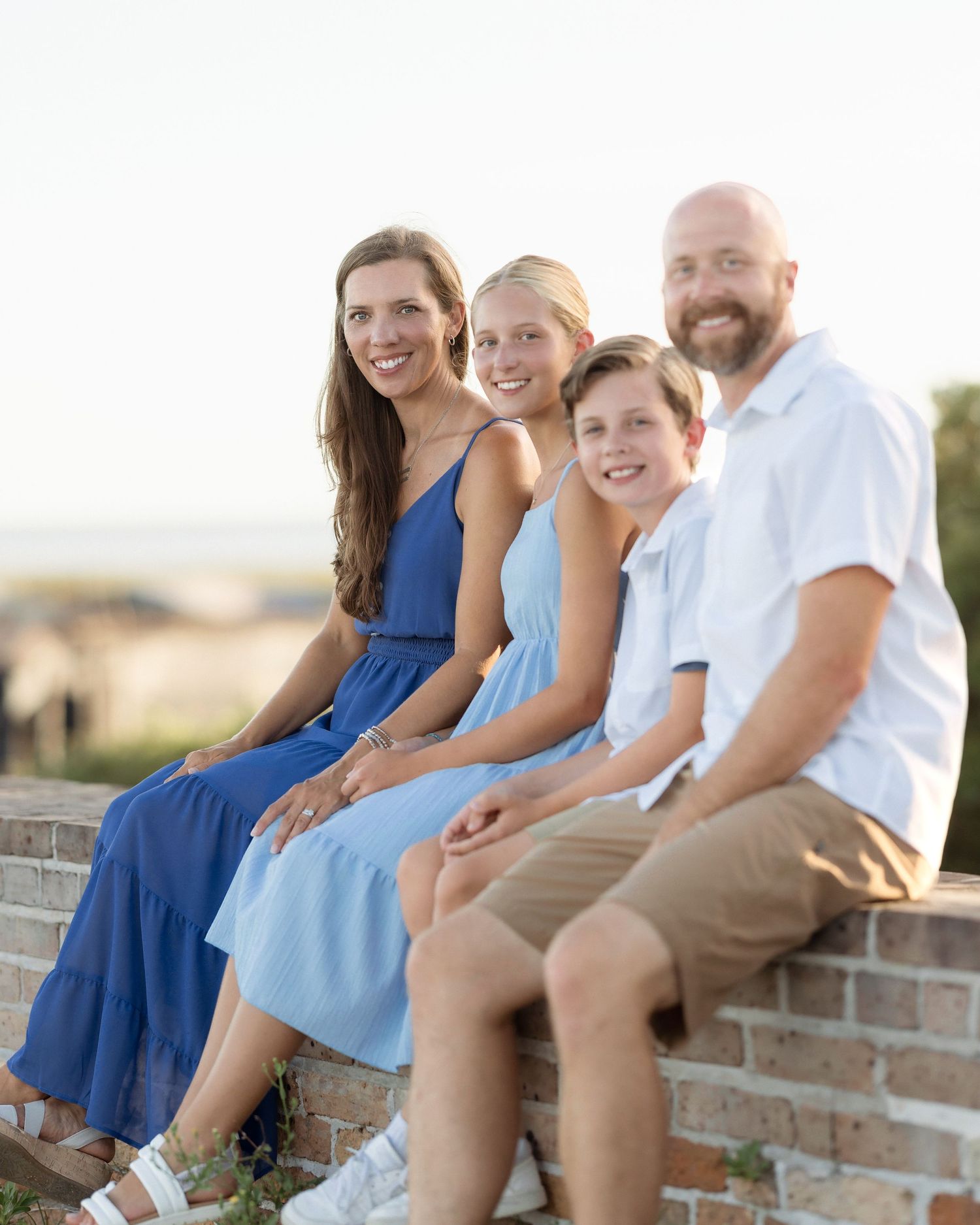 Family of four in blue outfits sitting on brick wall at beach during golden hour photo session.