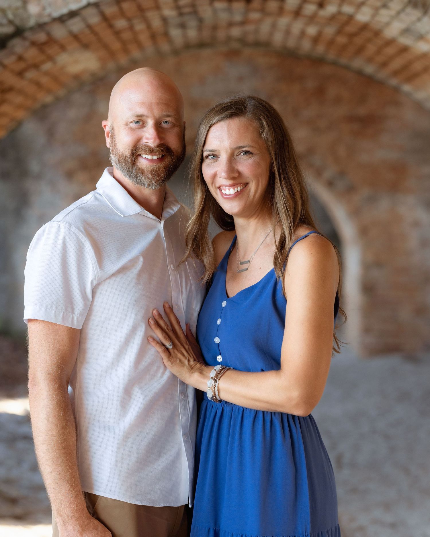 A smiling couple poses together under a brick archway, the man in a white shirt and the woman in a blue dress.