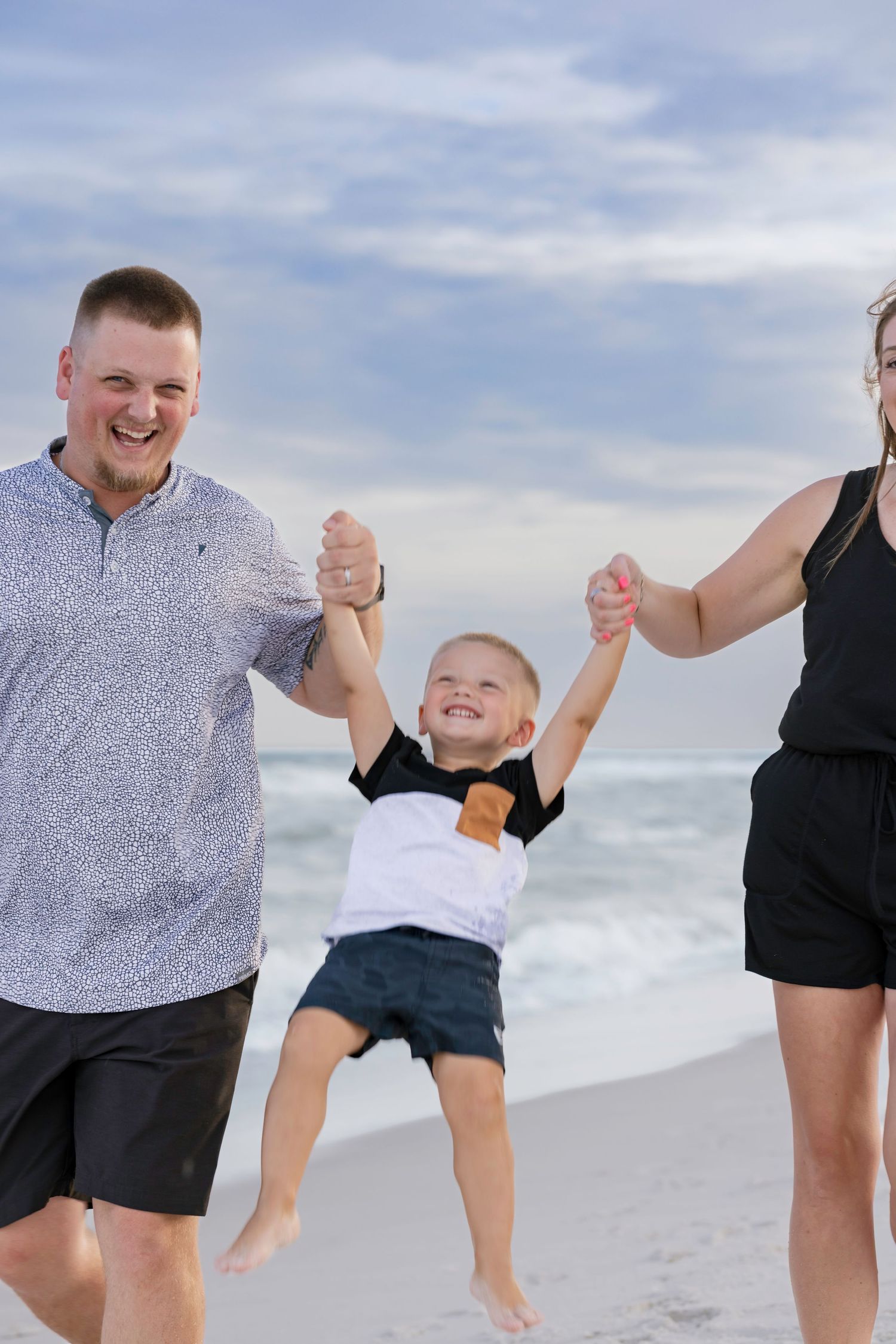 Happy family swings young boy by his hands on a sunny beach with ocean waves behind them.