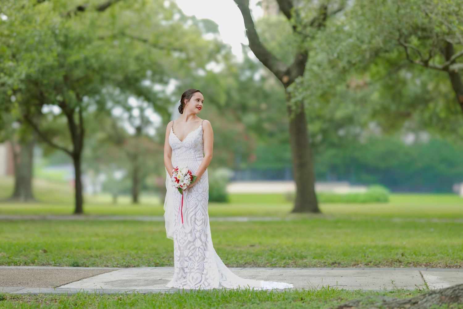 Bride in white lace gown holding bouquet stands on pathway surrounded by lush green trees in outdoor park setting.