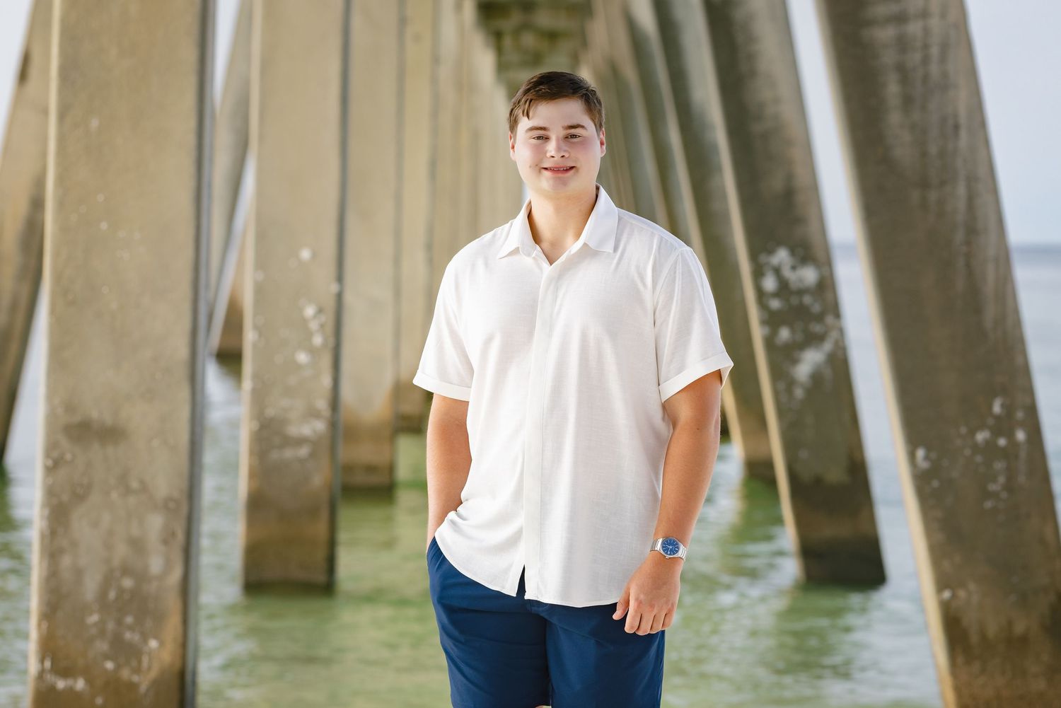 Young man in white shirt and blue pants poses under a pier with turquoise water in the background.