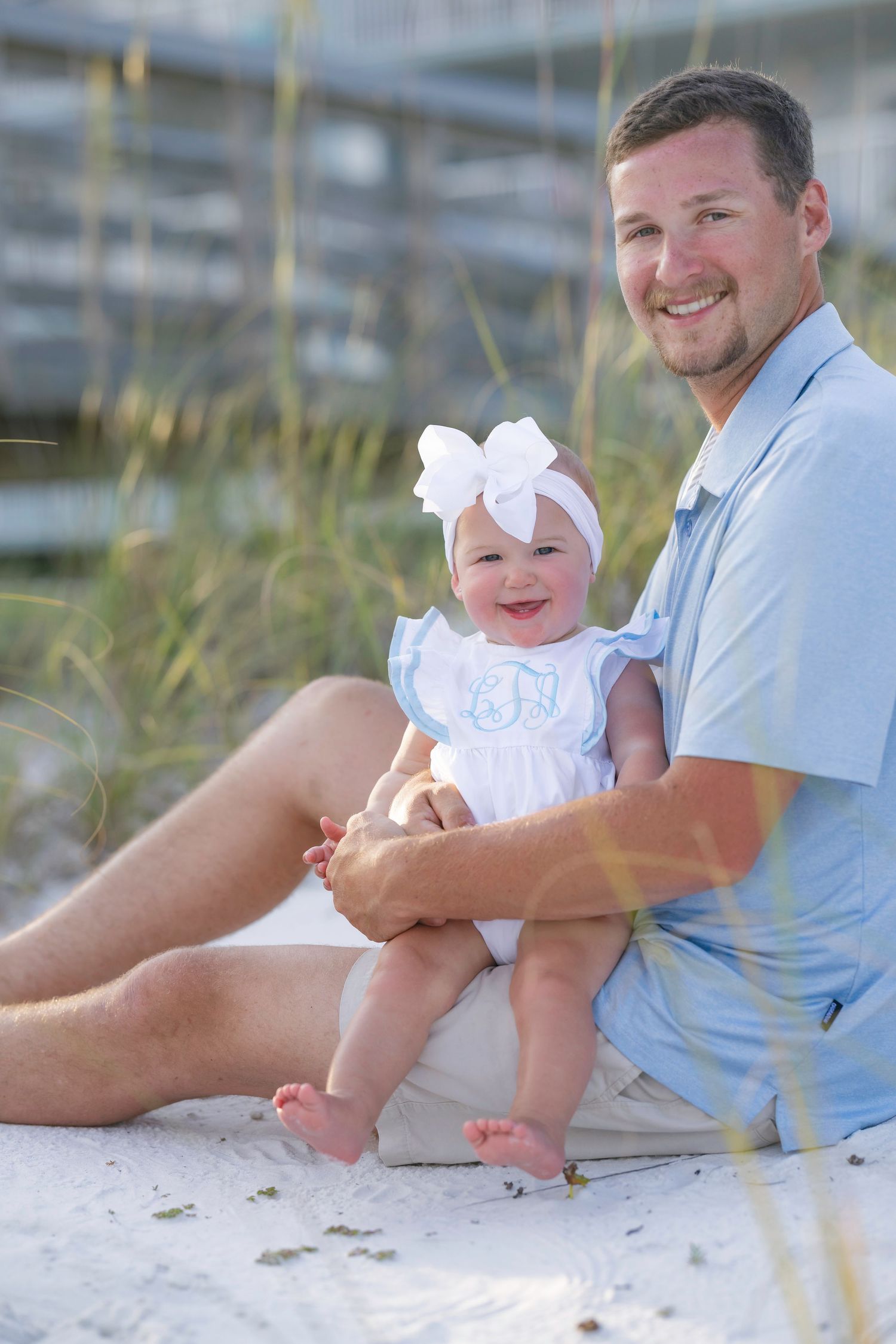 Smiling father sits on white sand beach holding happy baby girl in white monogrammed outfit with bow headband.