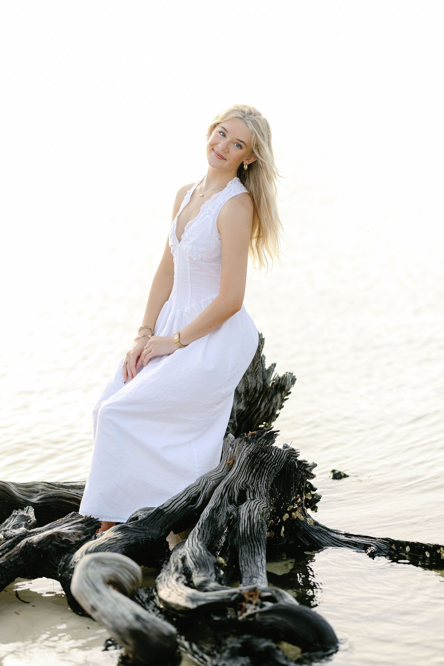 Young blonde woman in white dress sitting on driftwood by the water's edge in soft natural light.