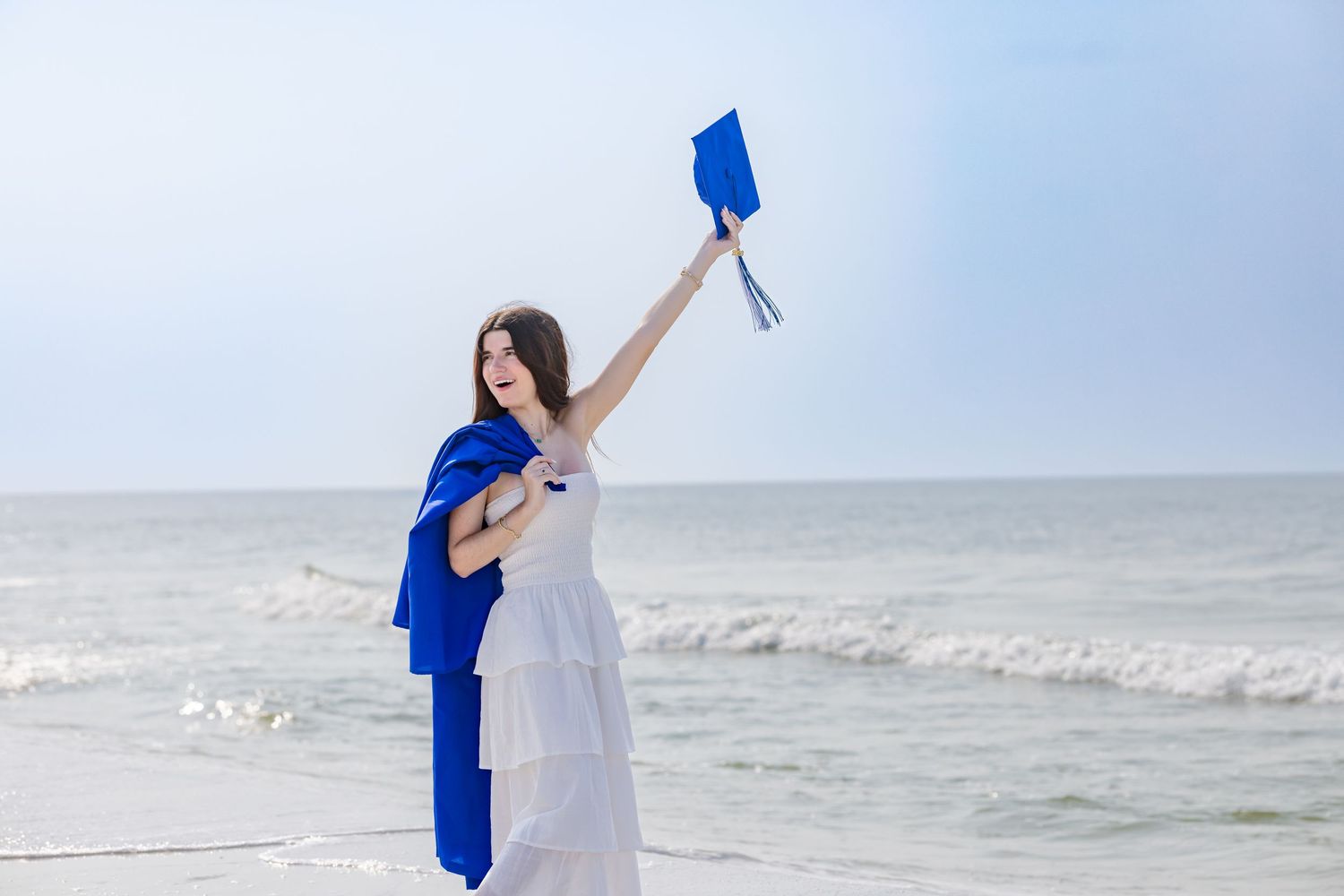 A graduating young woman in a white dress raises her blue cap triumphantly on a bright sunny beach.